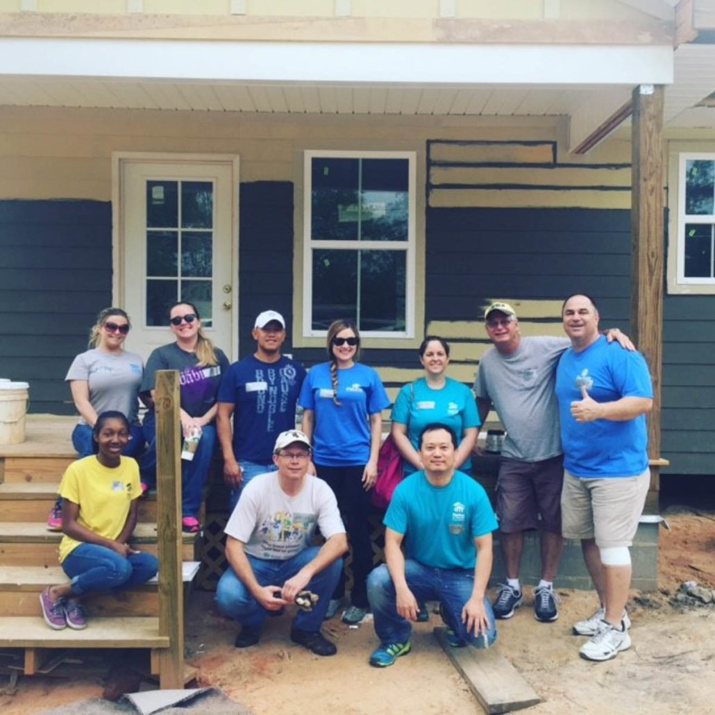 Group of nine people, including children and adults, posing in front of a partially built house with dark green siding, windows, and a white door, during a construction or volunteer project. Some are wearing casual clothes and sunglasses, with smiles