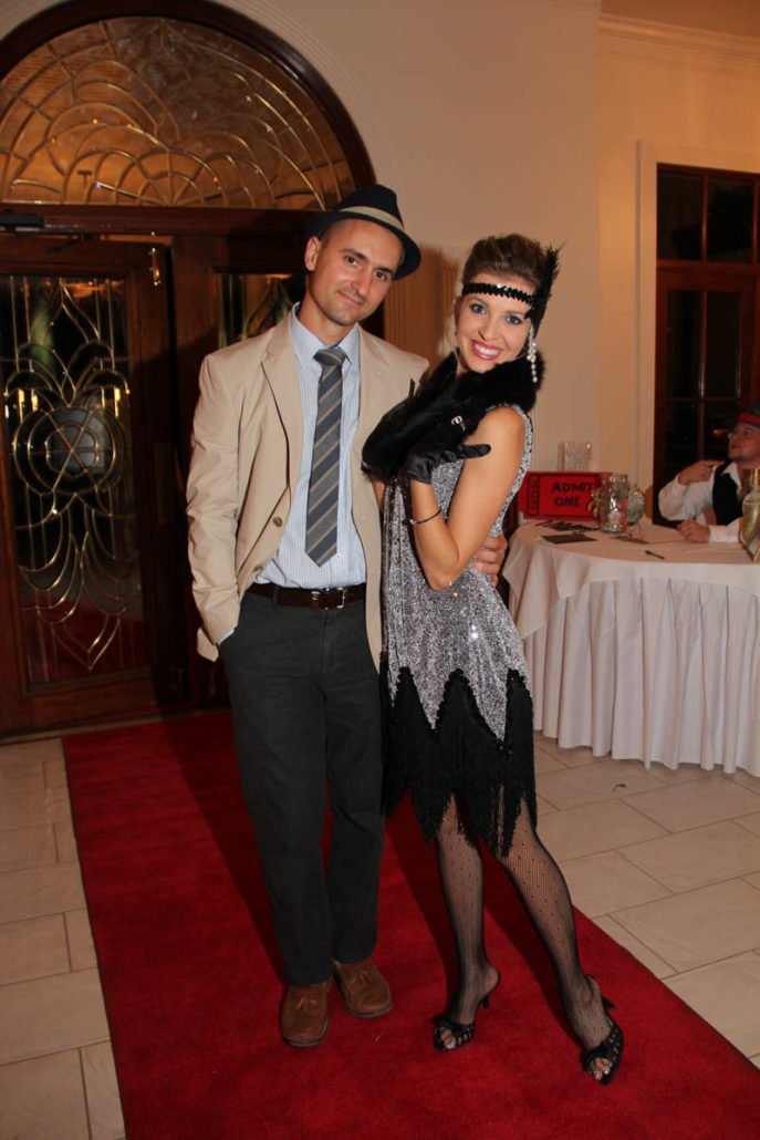A man and woman dressed in vintage 1920s attire standing on a red carpet at a themed party or event, with a table and a person sitting in the background.