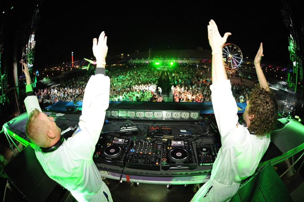 Two DJs performing on stage at a large outdoor music festival at night, with a crowd of people dancing and a ferris wheel in the background.