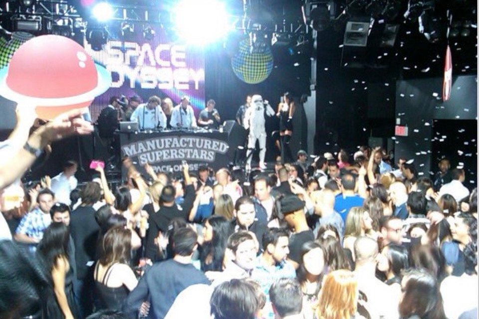 Crowd of people dancing and enjoying a concert at a large indoor venue with performers on stage and bright lights.