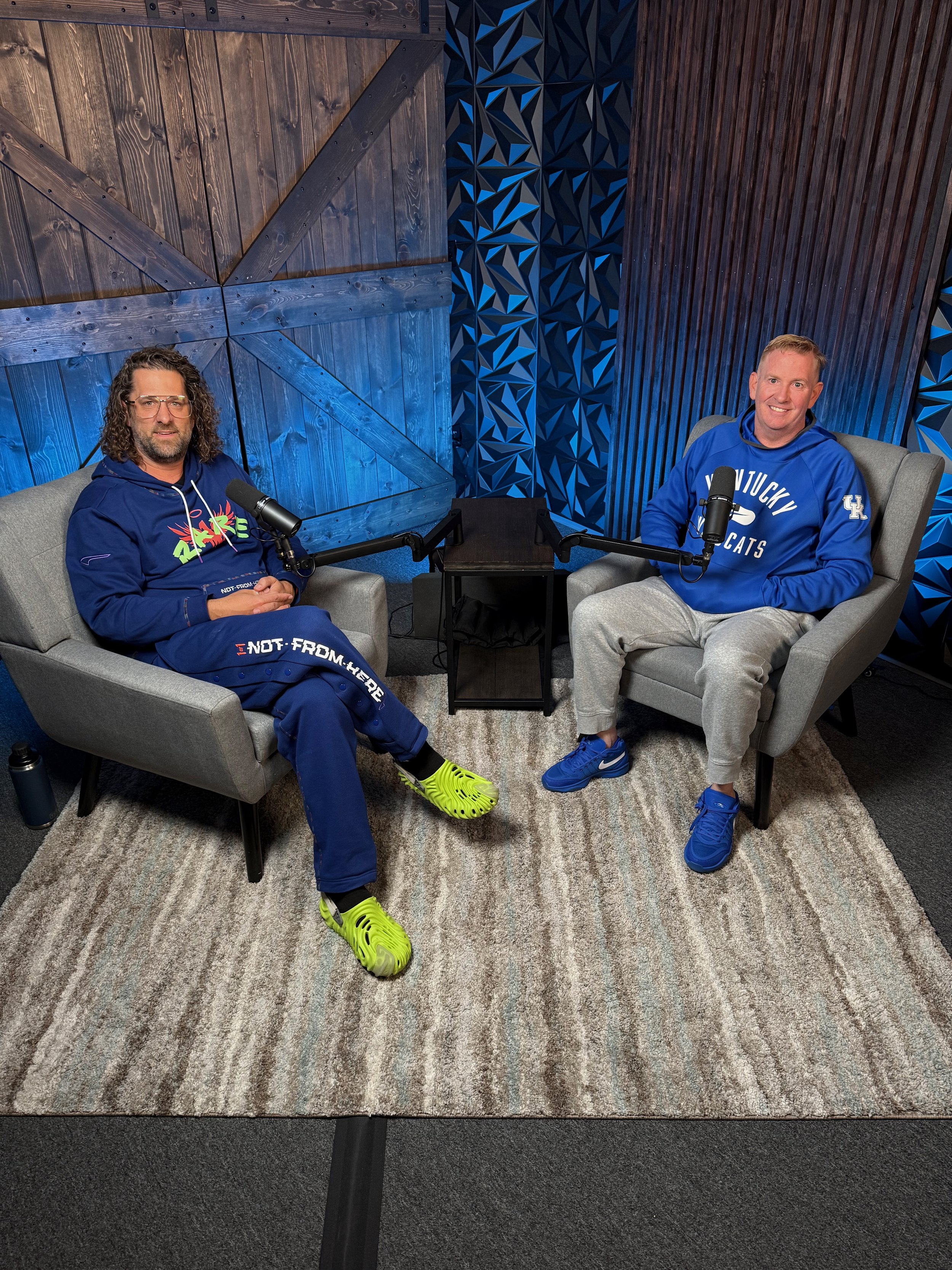 Two men sitting on gray armchairs in a podcast studio with microphones, wooden and blue patterned walls, and a small table between them.