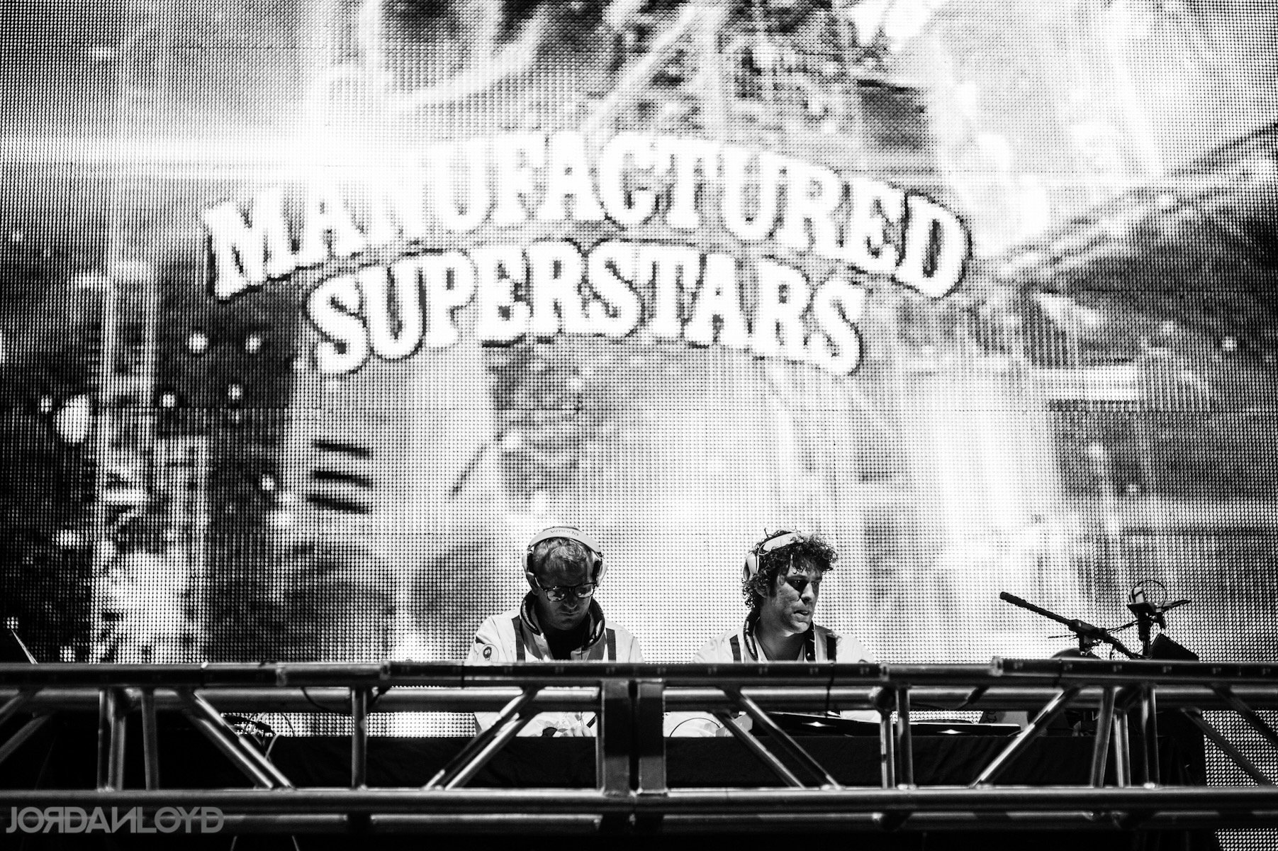 Two DJs wearing headphones and sunglasses perform behind a DJ booth with a large LED screen displaying the word "MANufactured SUPERSTARS" in the background.