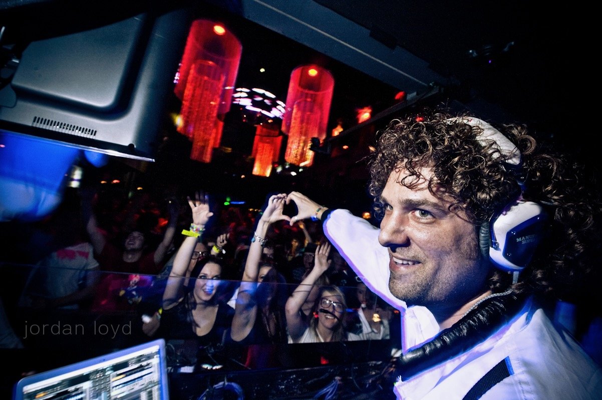 DJ with curly hair, wearing headphones, smiling, at a crowded nightclub with people dancing and colorful red pendant lights hanging from the ceiling.