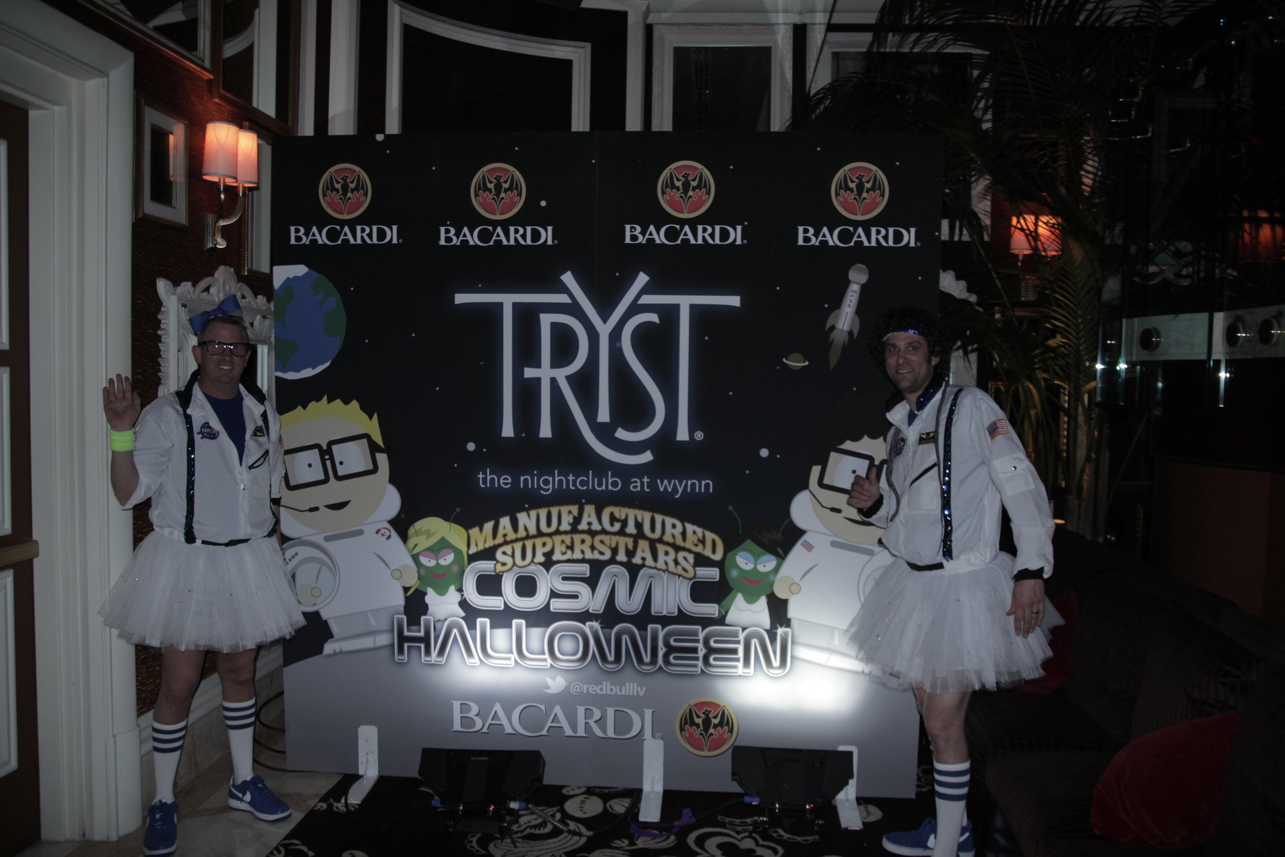 Two men dressed in astronaut costumes with tutus at a Halloween event, standing in front of a sign with space-themed graphics and event details.
