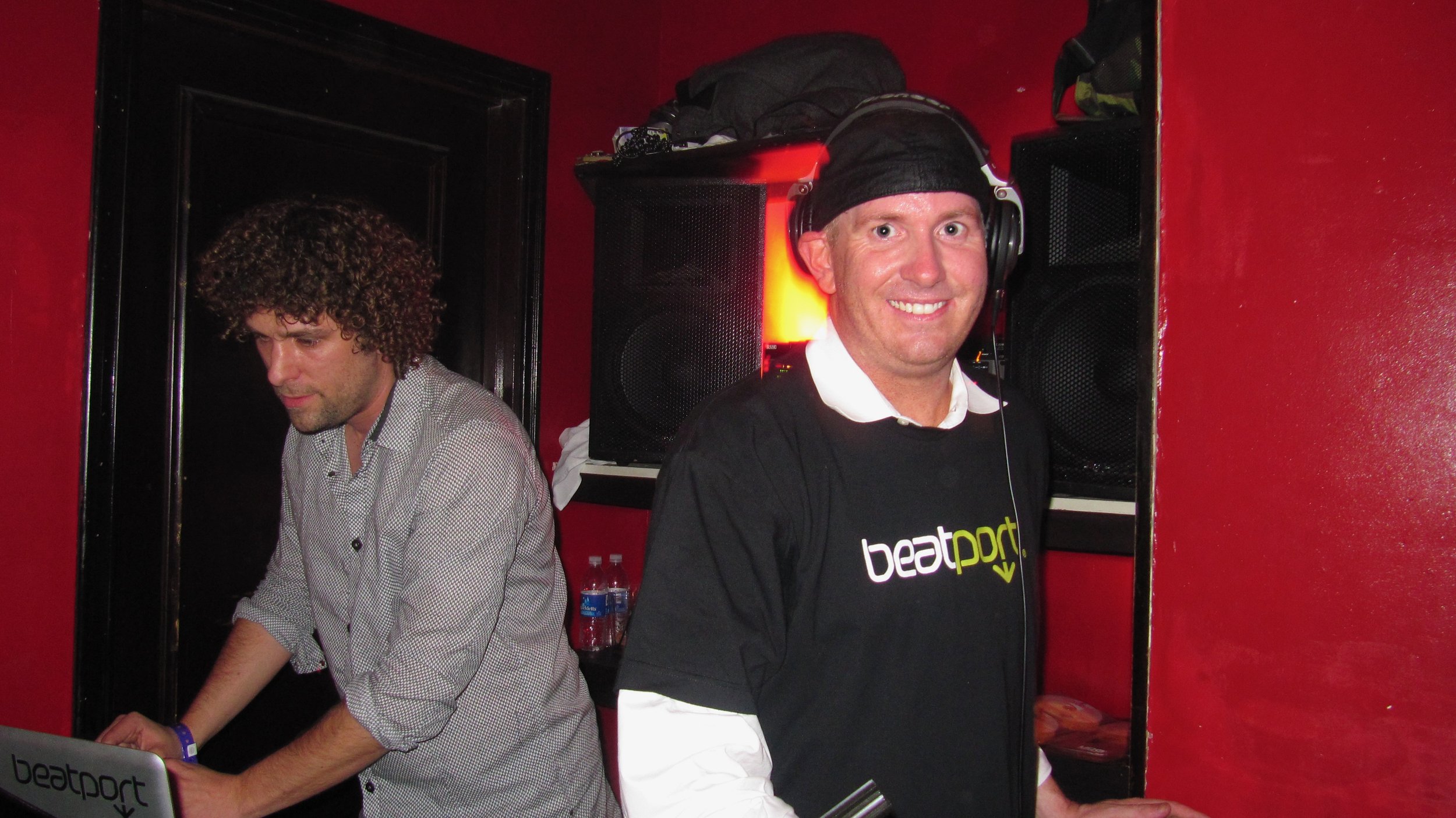 Two men at a DJ booth in a red-walled nightclub, one smiling wearing headphones and a black shirt with 'beatport' written on it, and the other focused on his equipment.