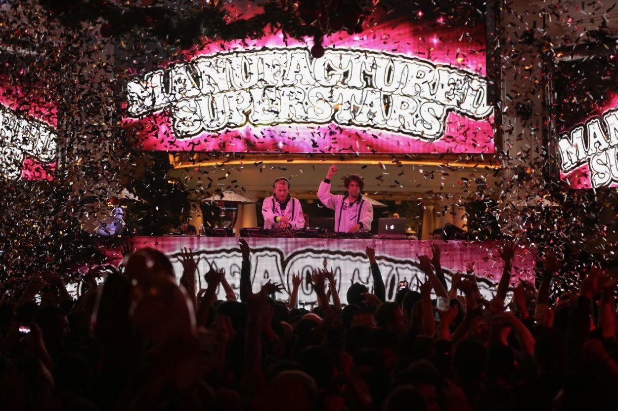 Two DJs perform on a stage at a music festival with a crowd dancing and cheering. Gold confetti falls from above, and bright pink neon signs display the festival's name, "Miami Ahoy! Ultra Miami 2023".