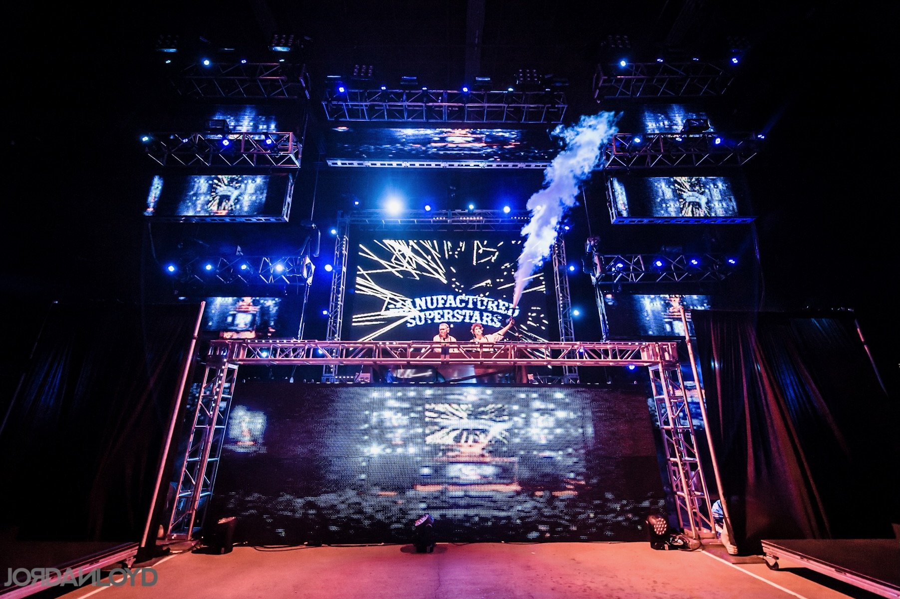 A stage set for a music performance with multiple large screens, blue and purple lighting, and a sign that says 'Manufactured Superstars' in the background.