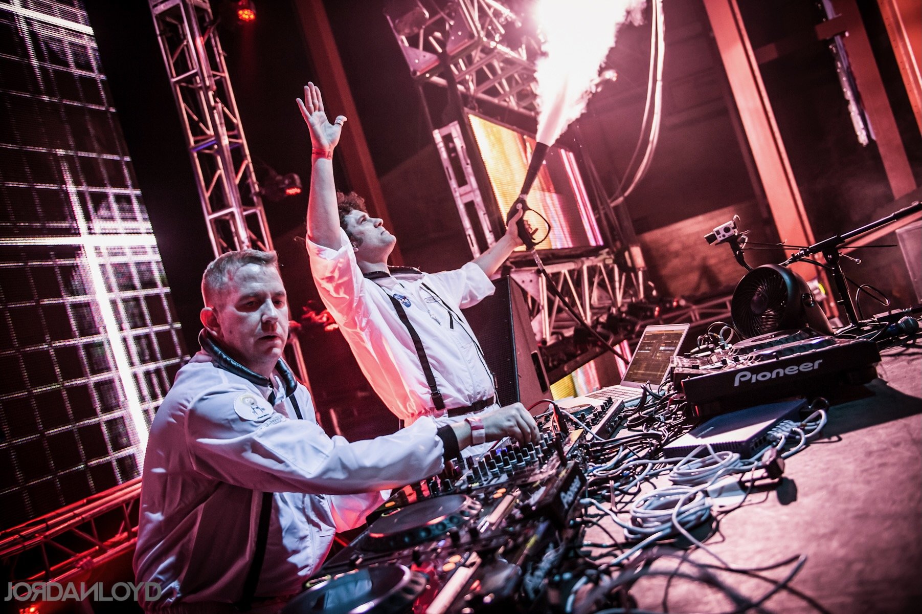 Two DJs in white jackets performing on stage with lighting and electronic equipment.