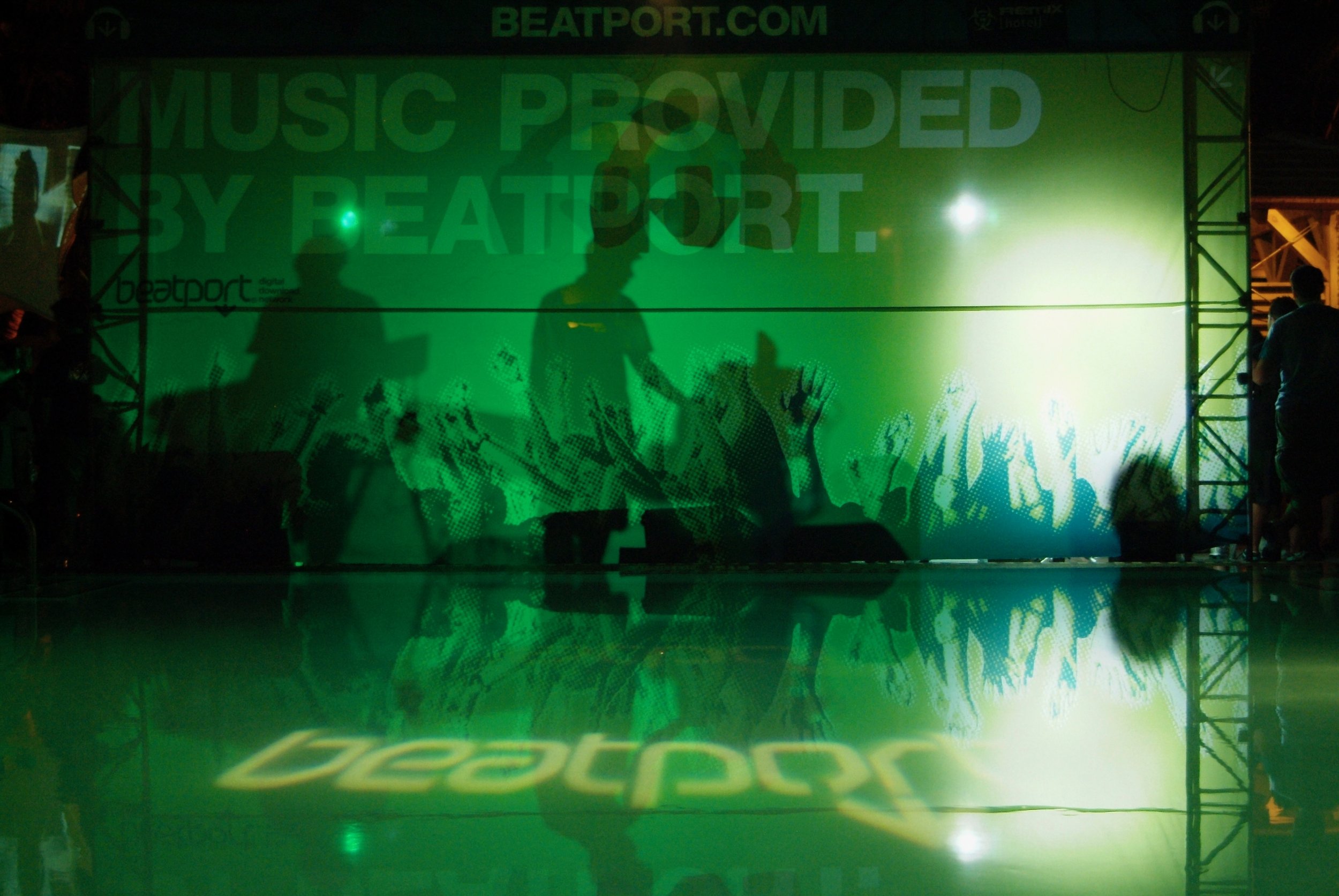 A large green screen at a Beatport event with silhouettes of people and raised hands, with the Beatport logo projected onto the floor and the backdrop, and a sign reading 'Music Provided by Beatport'