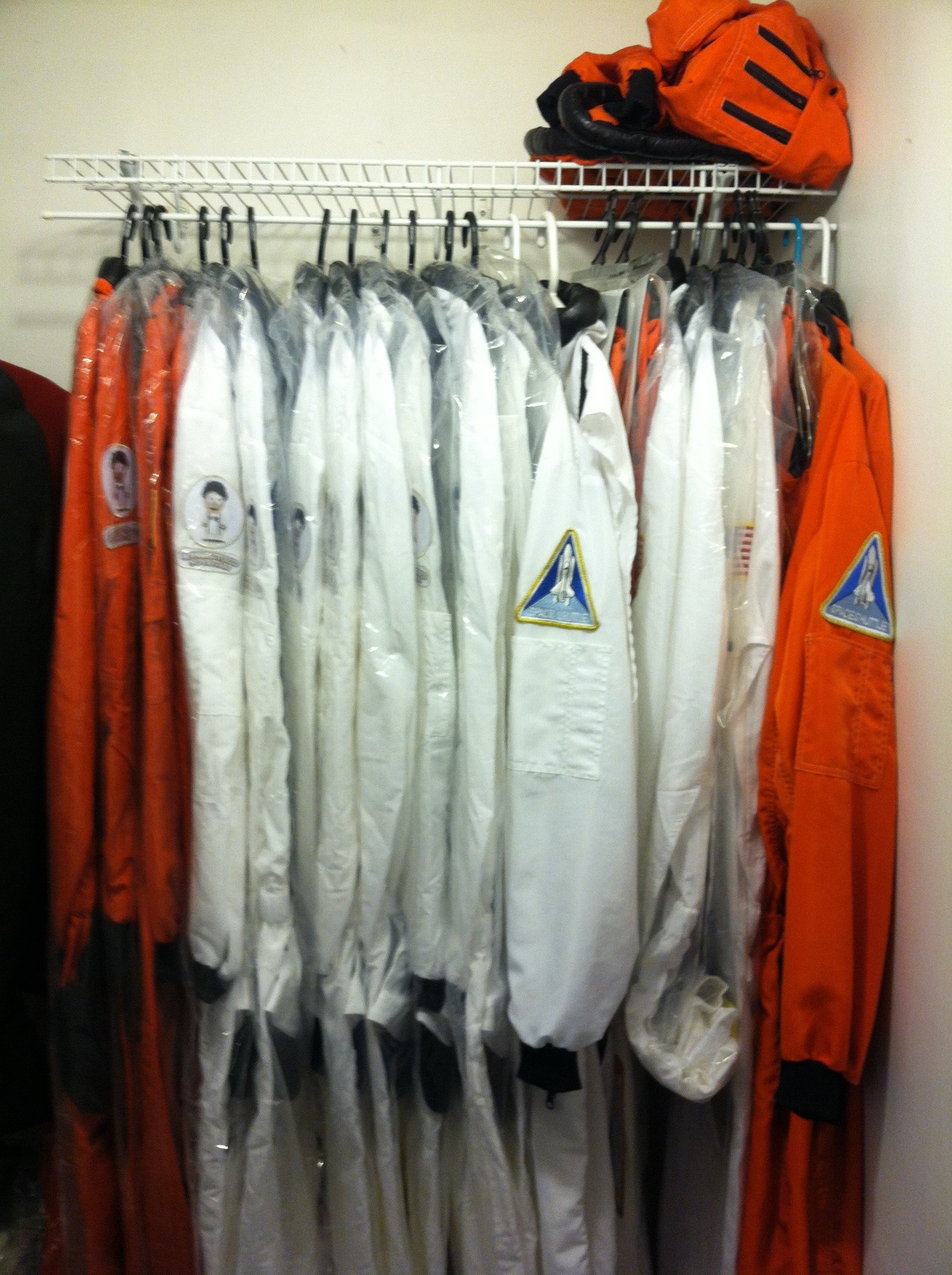 A closet with NASA astronaut suits, which are white and orange, hanging on a rack with some patches and symbols on the sleeves.