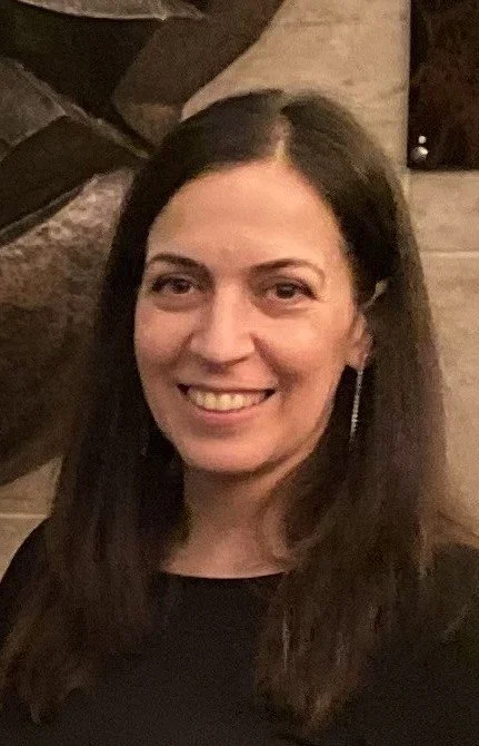 Smiling person with long dark hair wearing a black top against a neutral background.