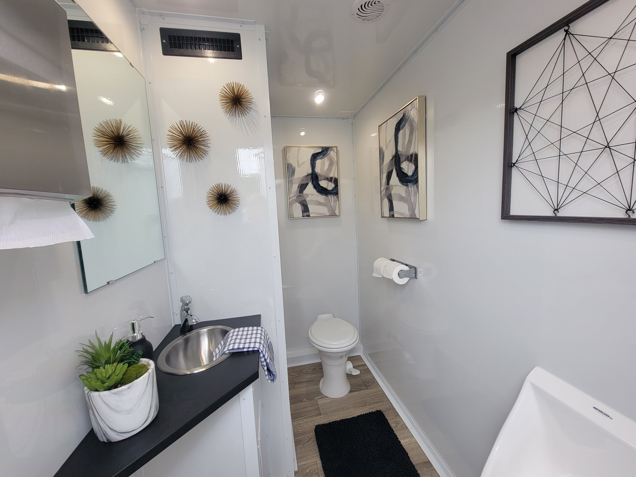 Modern bathroom with a sink, toilet, and urinal. The sink is set in a black countertop with a white and gray marble planter holding succulents. A small towel is draped over the sink edge. The wall is decorated with a mirror, abstract art, and metal w