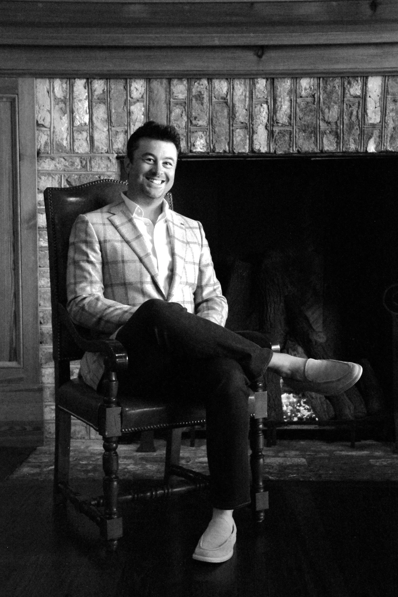 A man sitting in a chair in front of a brick fireplace, smiling, in a plaid blazer and dress shoes, in black and white.
