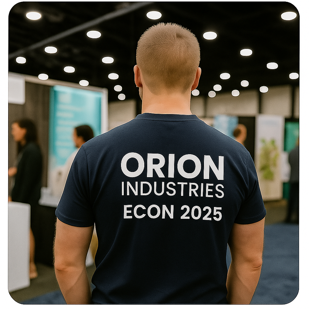 A person wearing a navy blue T-shirt with white text reading 'ORION INDUSTRIES ECON 2025' on the back, standing at an indoor conference or exhibition with blurred background of displays and other attendees.