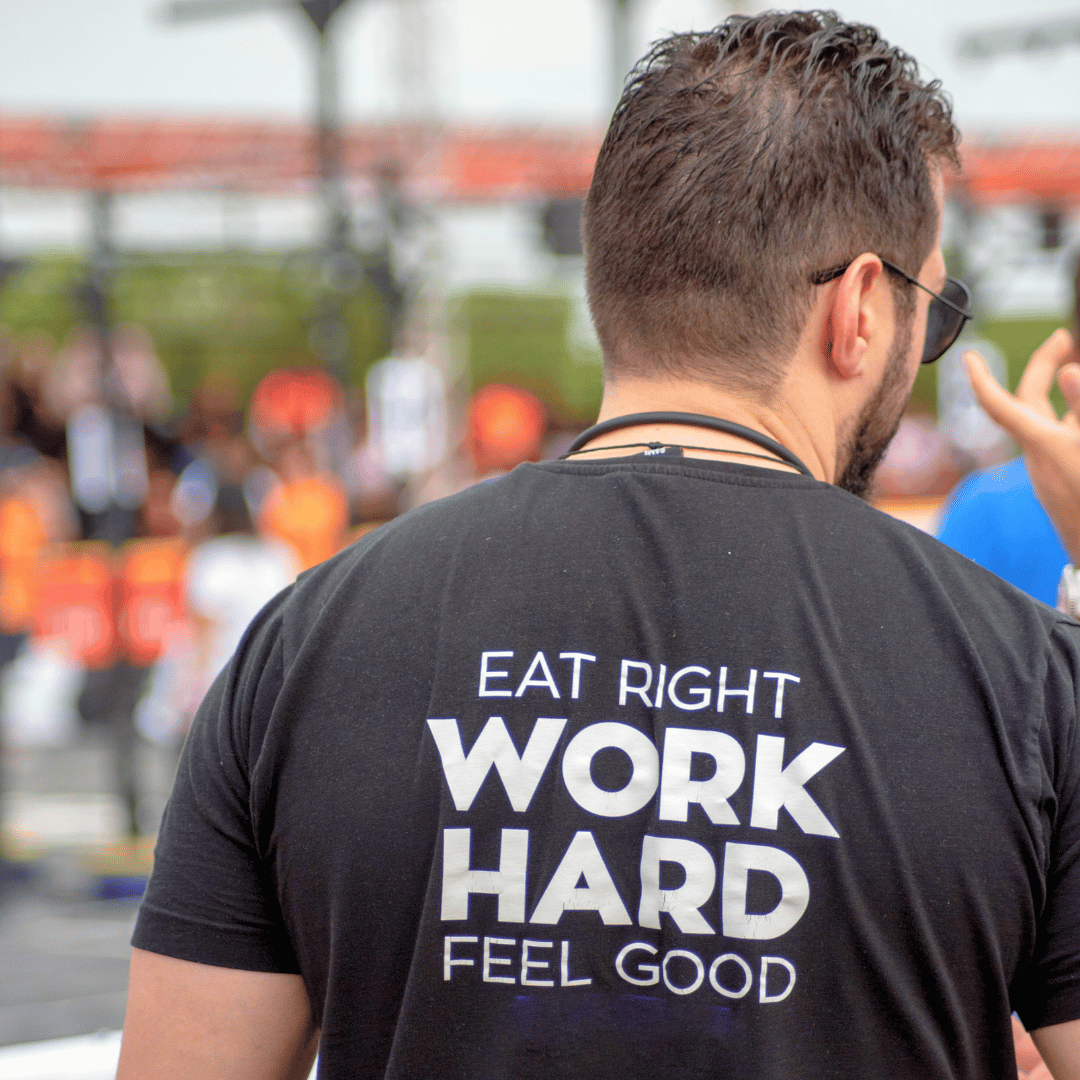 Back of a man wearing a black T-shirt with the text "Eat right, work hard, feel good" printed on it. The background shows a blurred outdoor scene with people and structures.