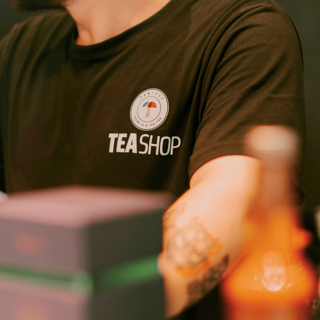 Close-up of a person wearing a black T-shirt with a logo for TEA SHOP, with the words "TEA SHOP" printed on it. The person has tattoos on their arm and is in a setting that appears to be a tea shop or cafe.
