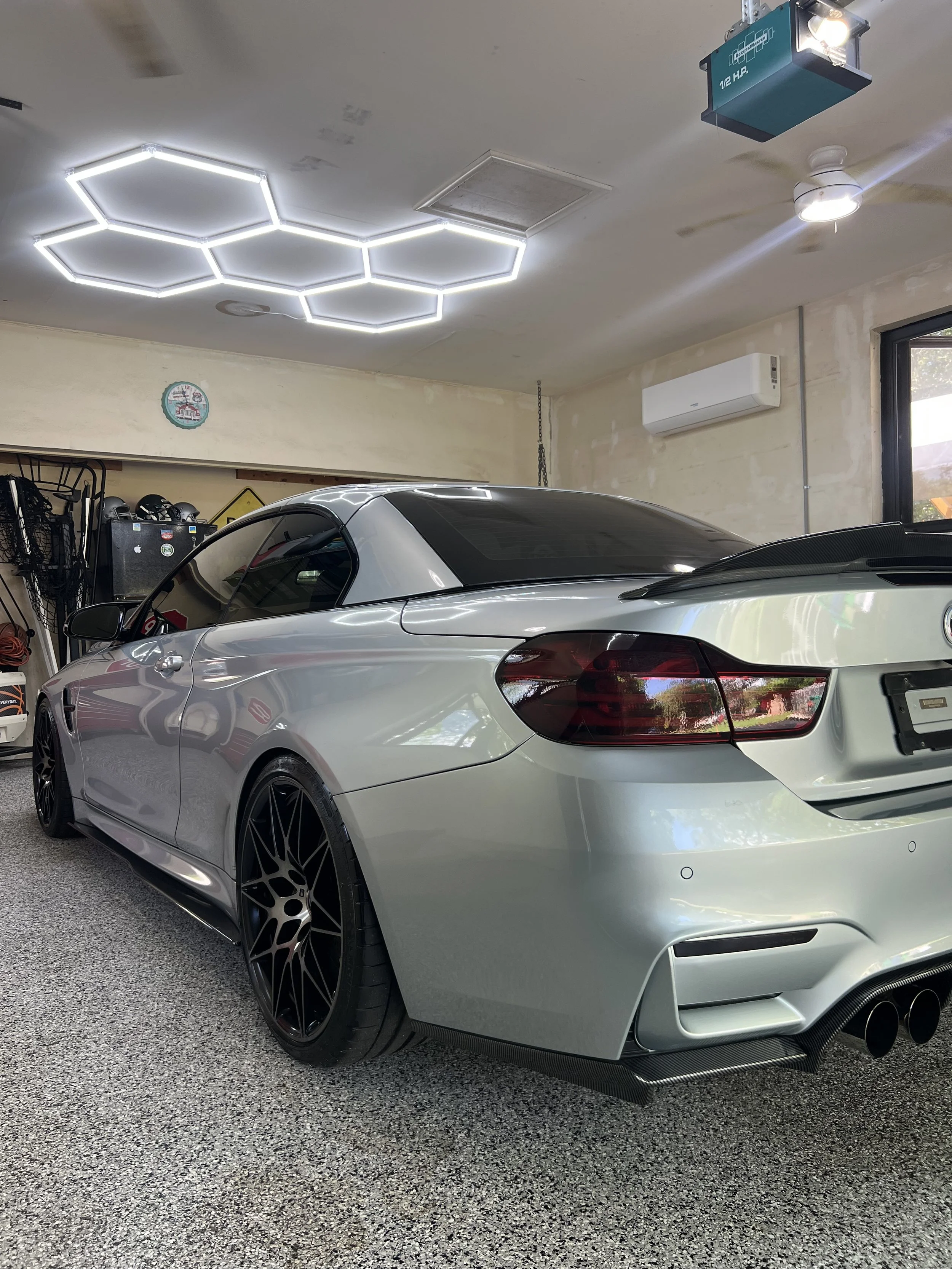 A silver sports car with black rims parked in a garage, with hexagon-shaped LED ceiling lights and wall-mounted storage and tools.