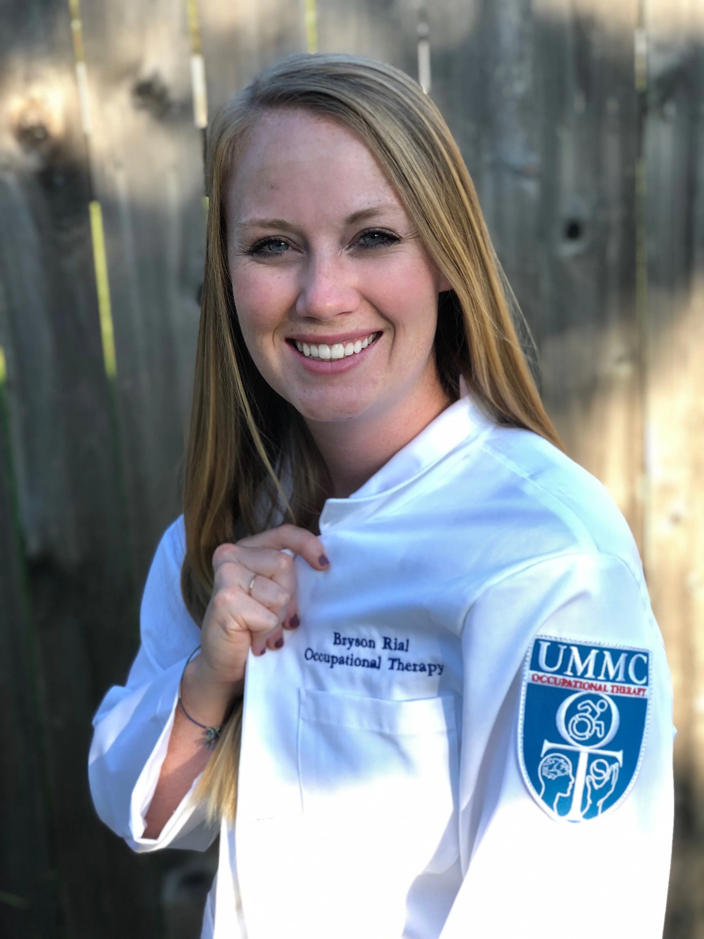 A smiling woman with long red hair in occupational therapy attire, wearing a white coat with her name 'Bryson Rial' and 'Occupational Therapy' embroidered on it, standing outdoors in front of a wooden fence.