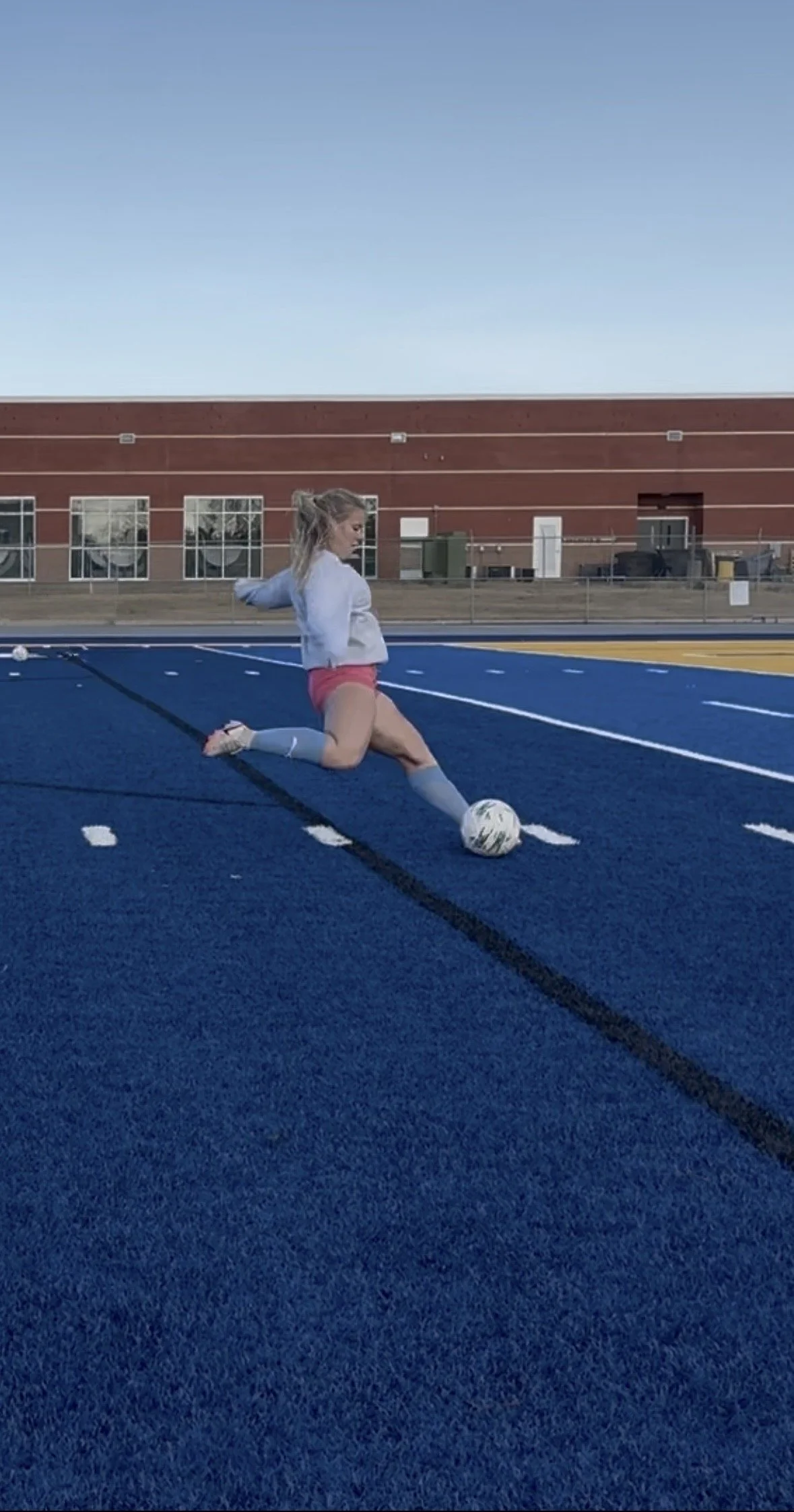 A young woman playing soccer on a blue artificial turf field outside with a brick building in the background, kicking a white soccer ball.