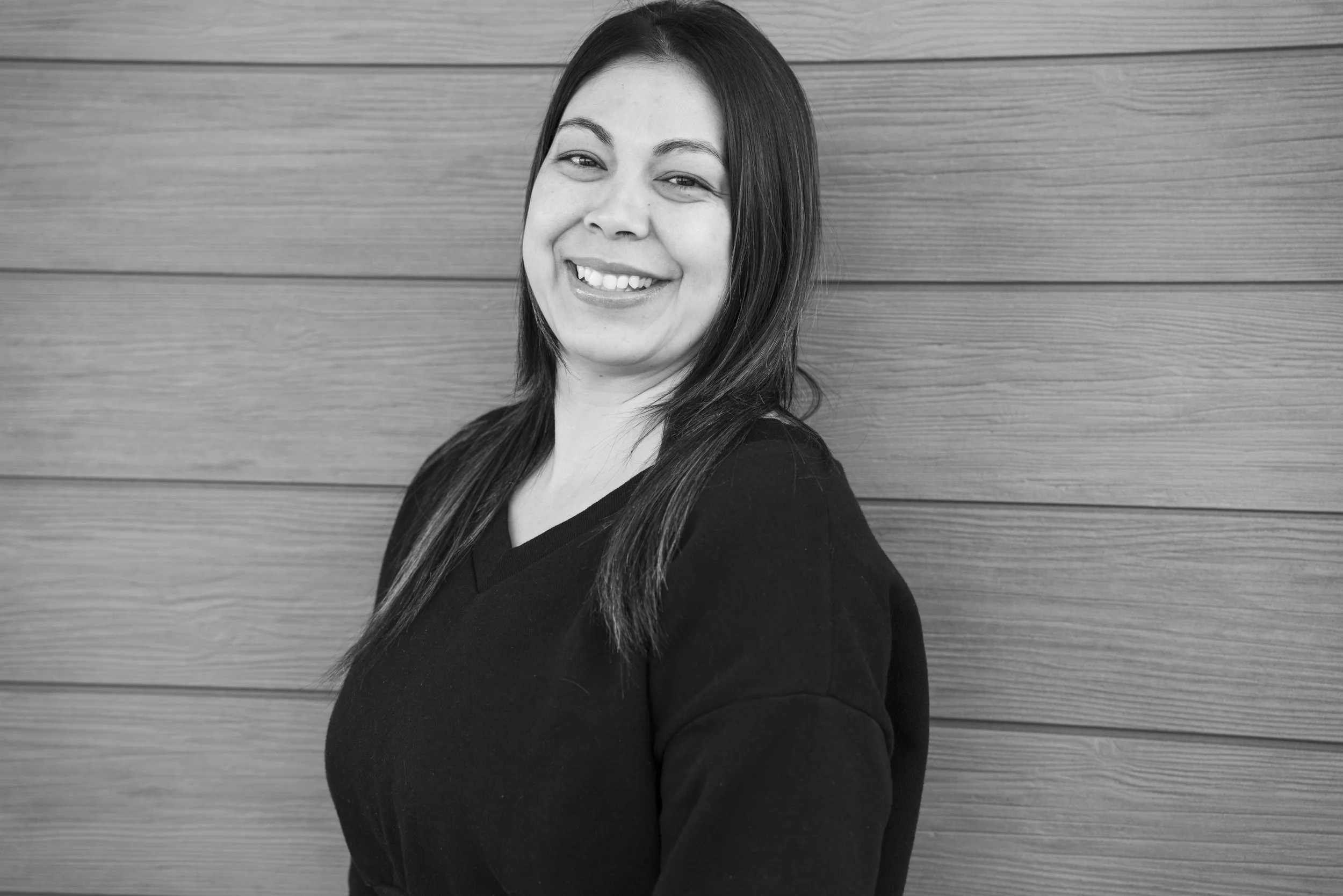 Black and white portrait of a smiling woman with long dark hair, wearing a black top, standing against a wooden wall.