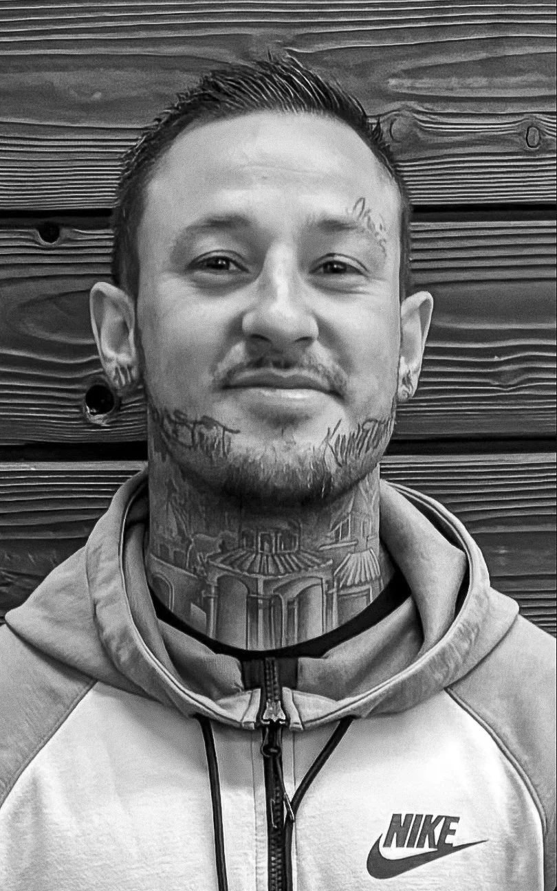 Man with facial and neck tattoos wearing a gray Nike hoodie, standing against a dark wooden background.