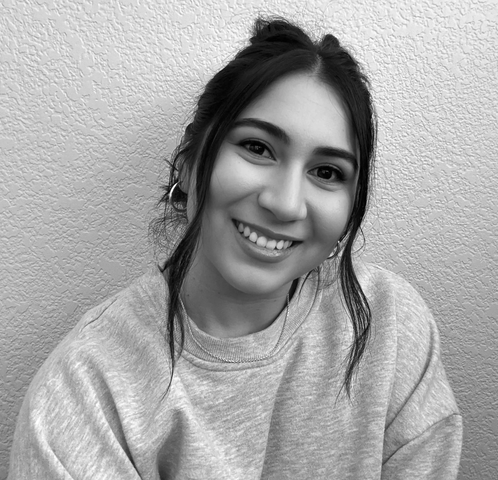 Black and white photo of a young woman smiling, wearing a sweatshirt and hoop earrings, in front of a textured wall.