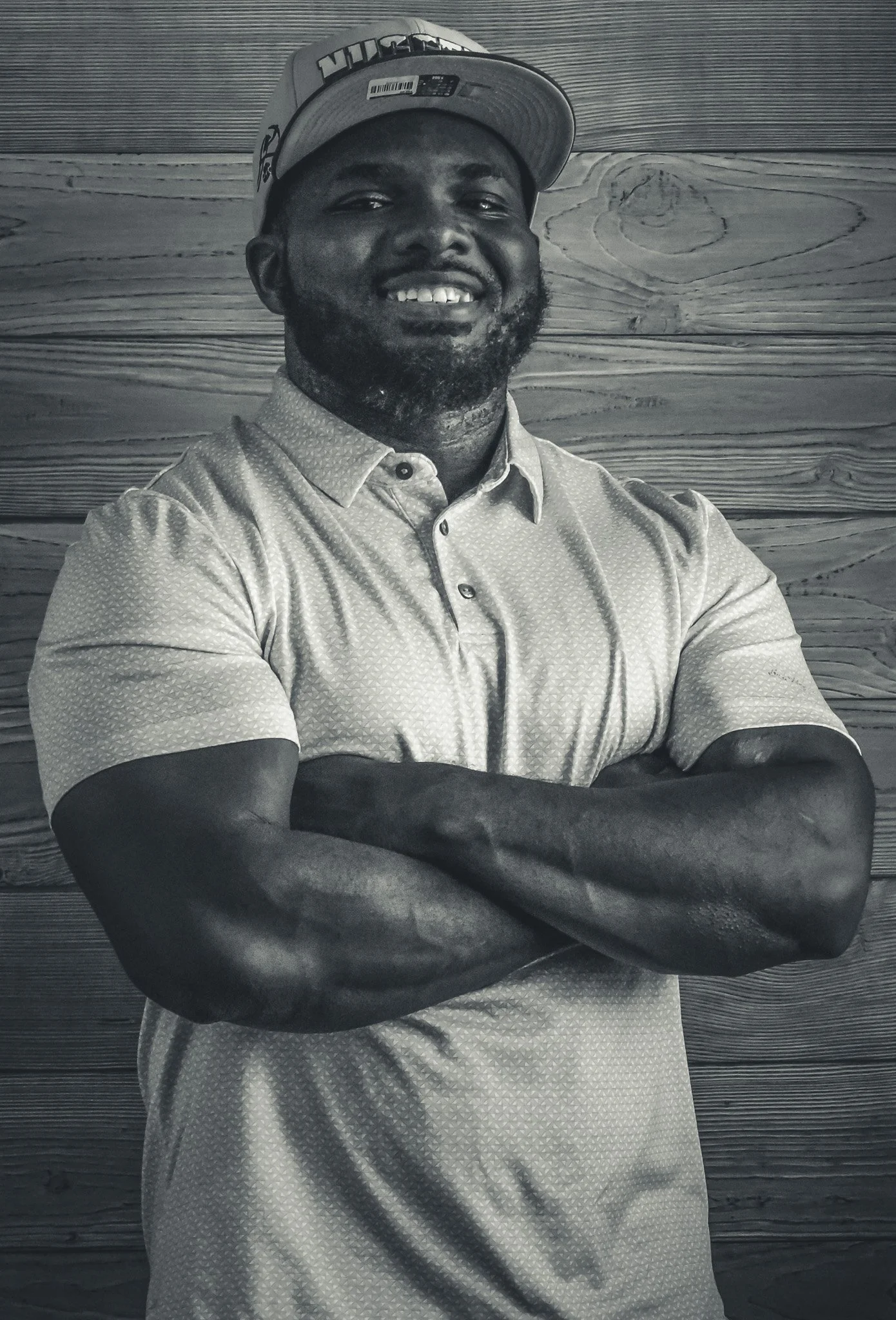 A man smiling with arms crossed, wearing a baseball cap and a patterned button-up shirt, standing in front of a wooden wall.