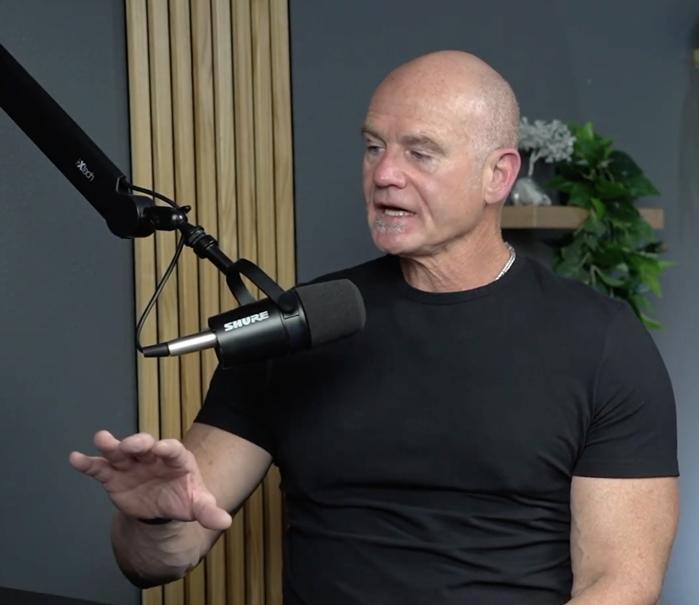 A man with a shaved head and black t-shirt speaking into a microphone during a recording or interview in a studio setting with a wooden panel and plant in the background.