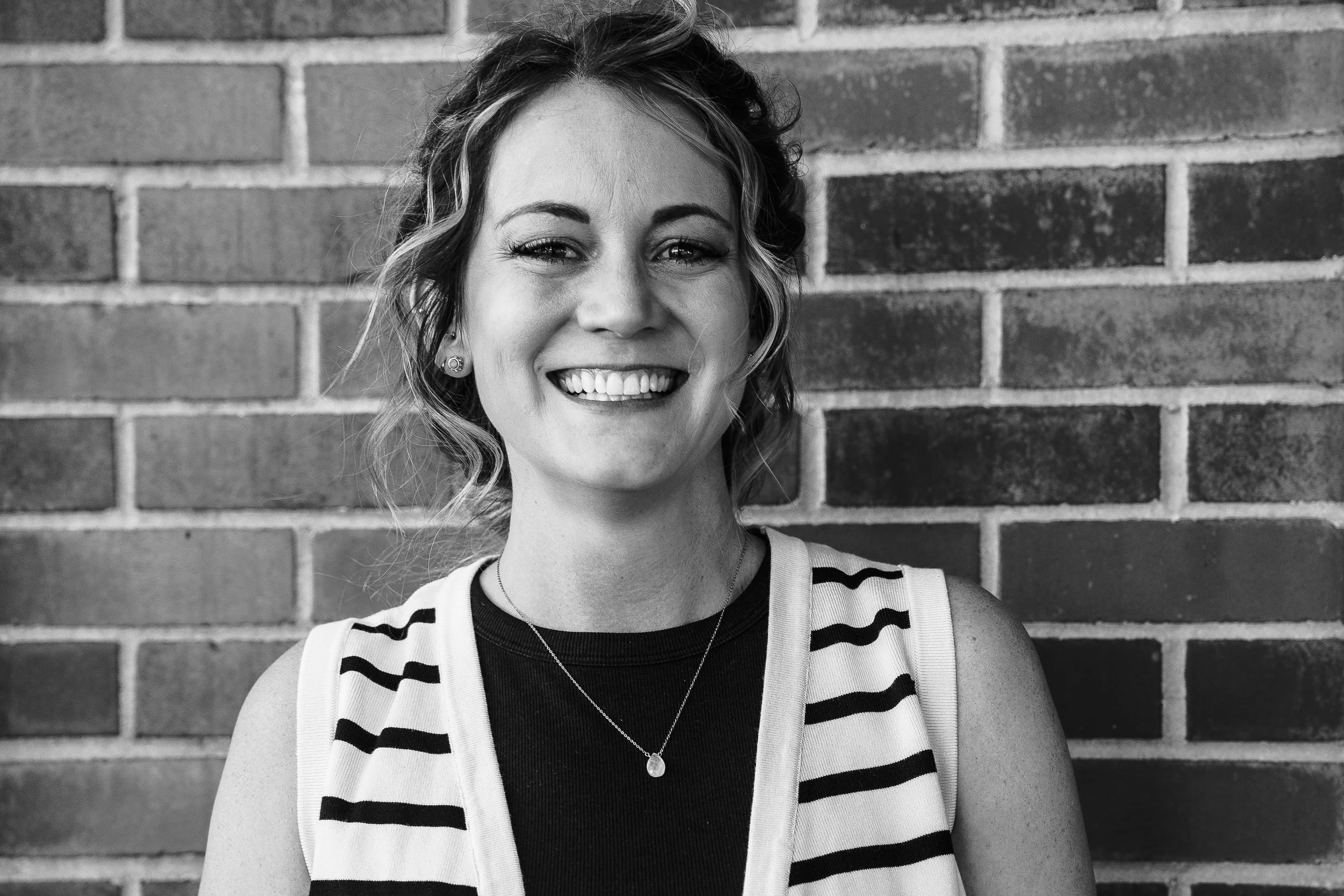 A woman with blonde, wavy hair smiling outdoors next to a calm body of water with red rock cliffs on one side and a cloudy sky overhead.
