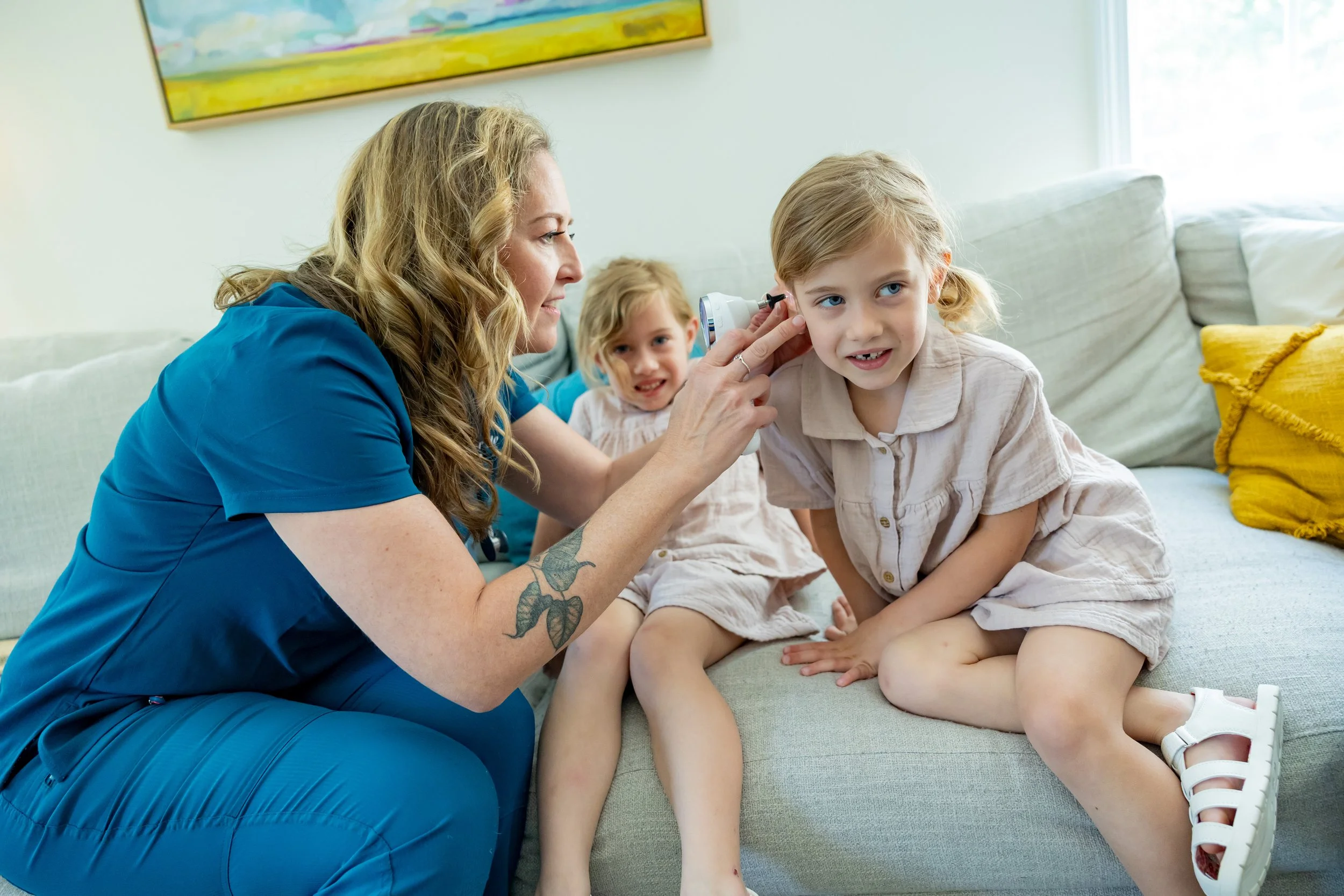 A pediatrician with long curly blonde hair, wearing blue scrubs, is examining a young girl's ear with an otoscope, while two other young girls sit beside her on a couch, watching.