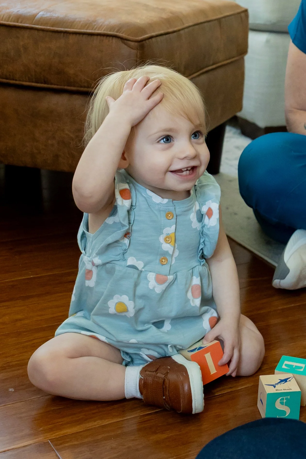 A young girl with blonde hair, blue eyes, wearing a blue floral dress, brown shoes, sitting on a wooden floor, smiling, with her hand on her head, surrounded by alphabet blocks.