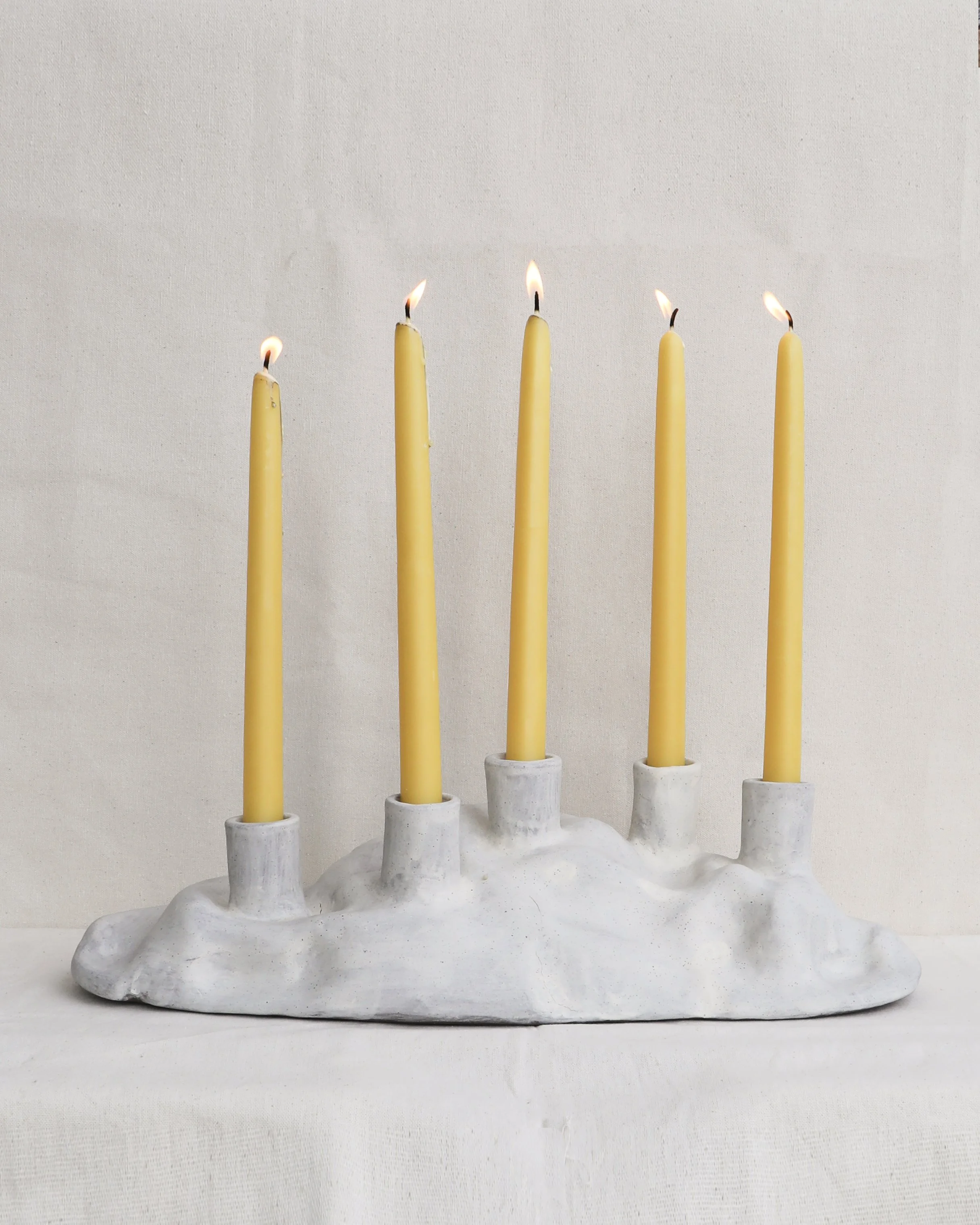 A white candle holder with multiple indented sections supports five yellow candles in a row, some of which are lit, set against a plain, light-colored background.