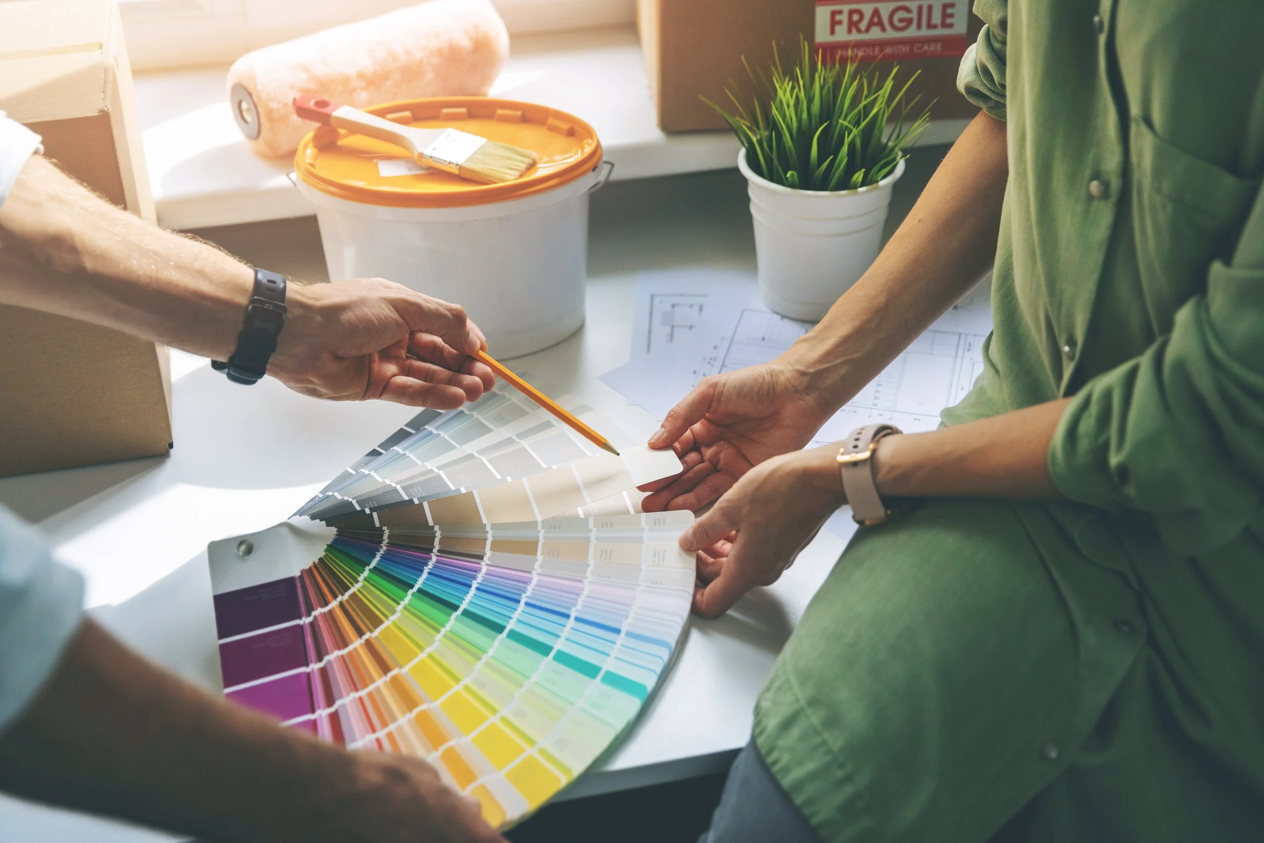 Two people discussing a color palette and paint sample swatch, with paint project plans and a potted plant in the background.