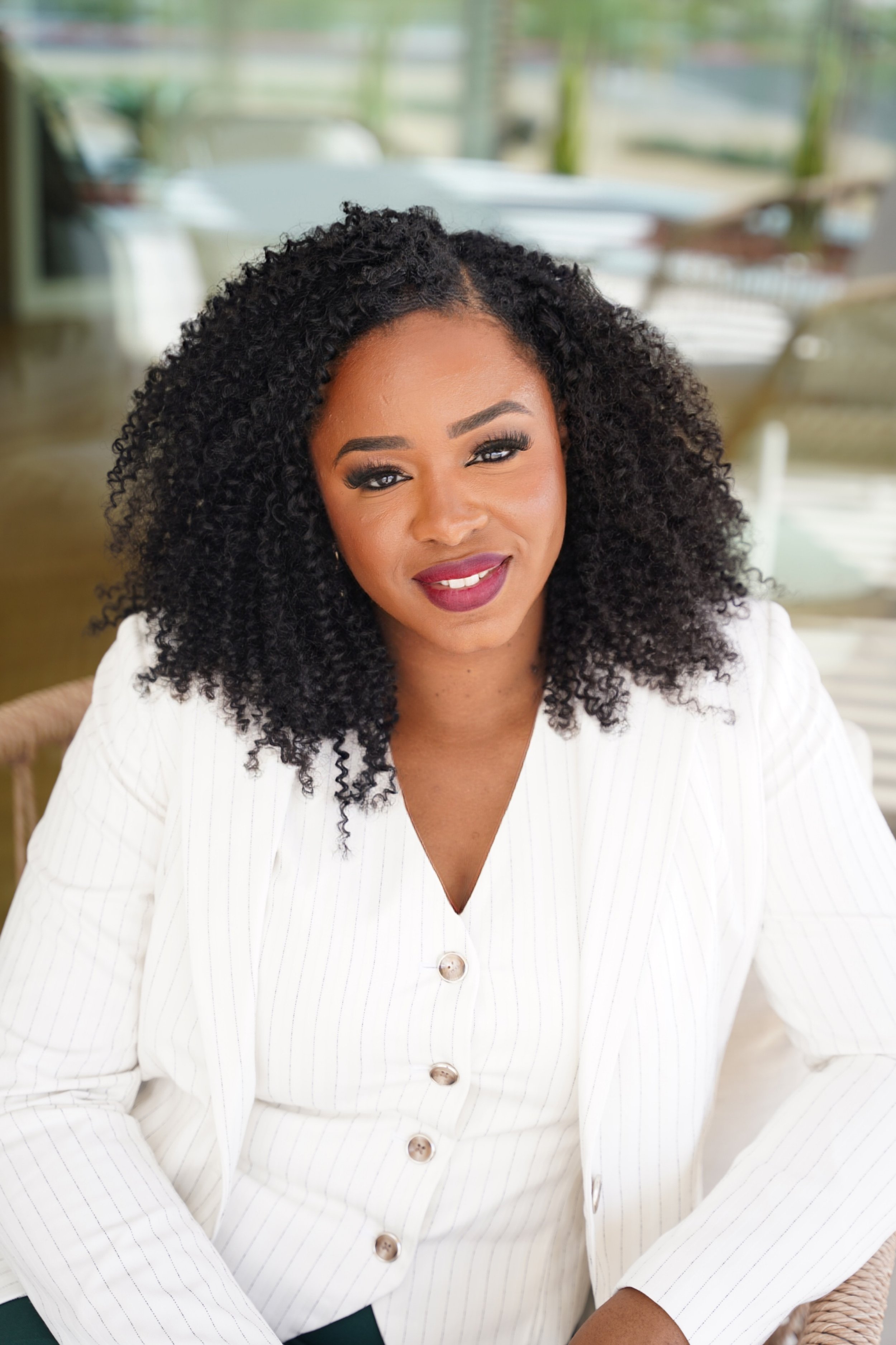 Portrait of a woman with curly black hair wearing a white pinstripe blazer, smiling softly, seated indoors with blurred background.