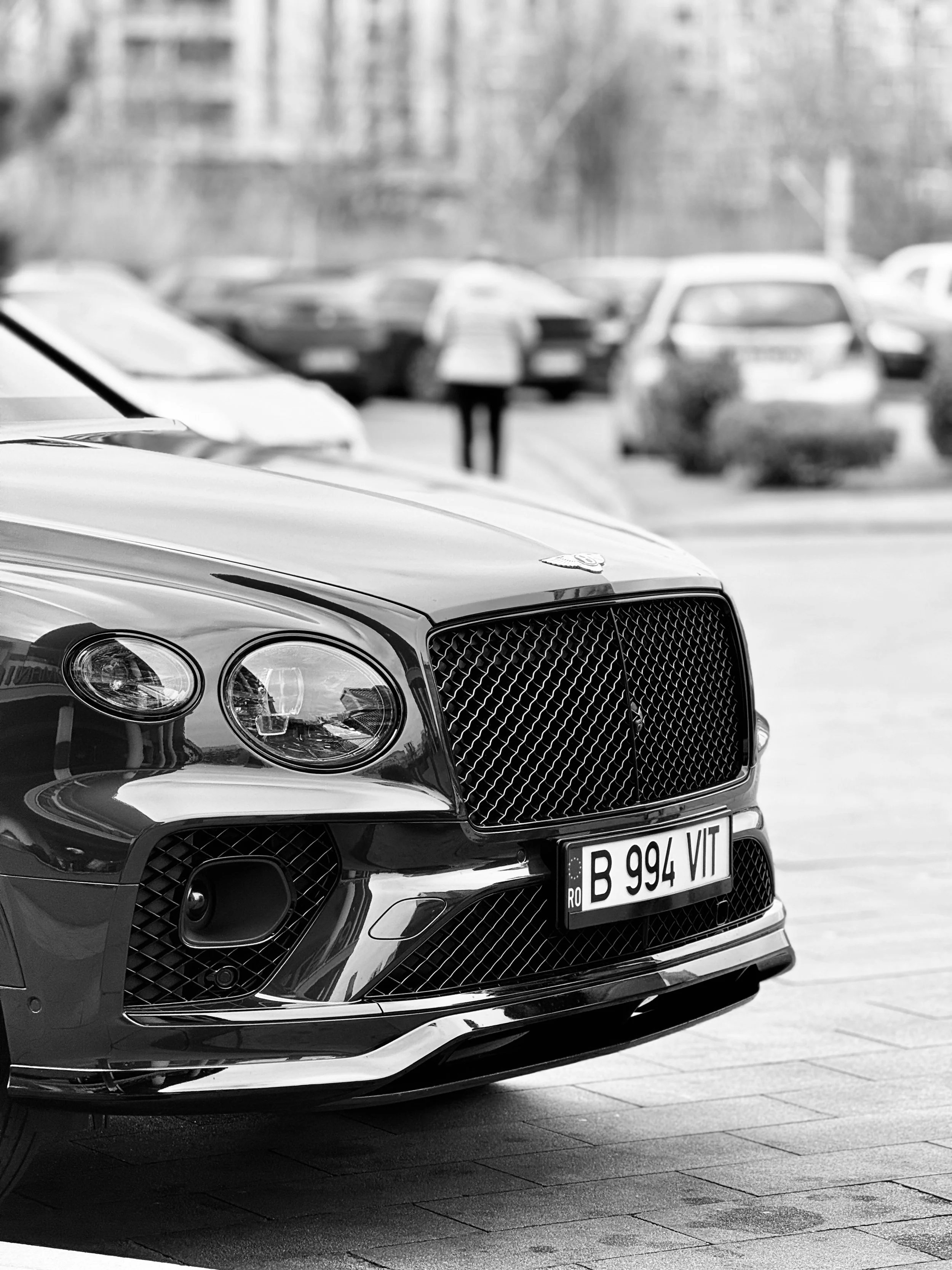 Close-up of a luxury car with a distinctive grille and headlights, parked in a lot with other vehicles in the background, in black and white.