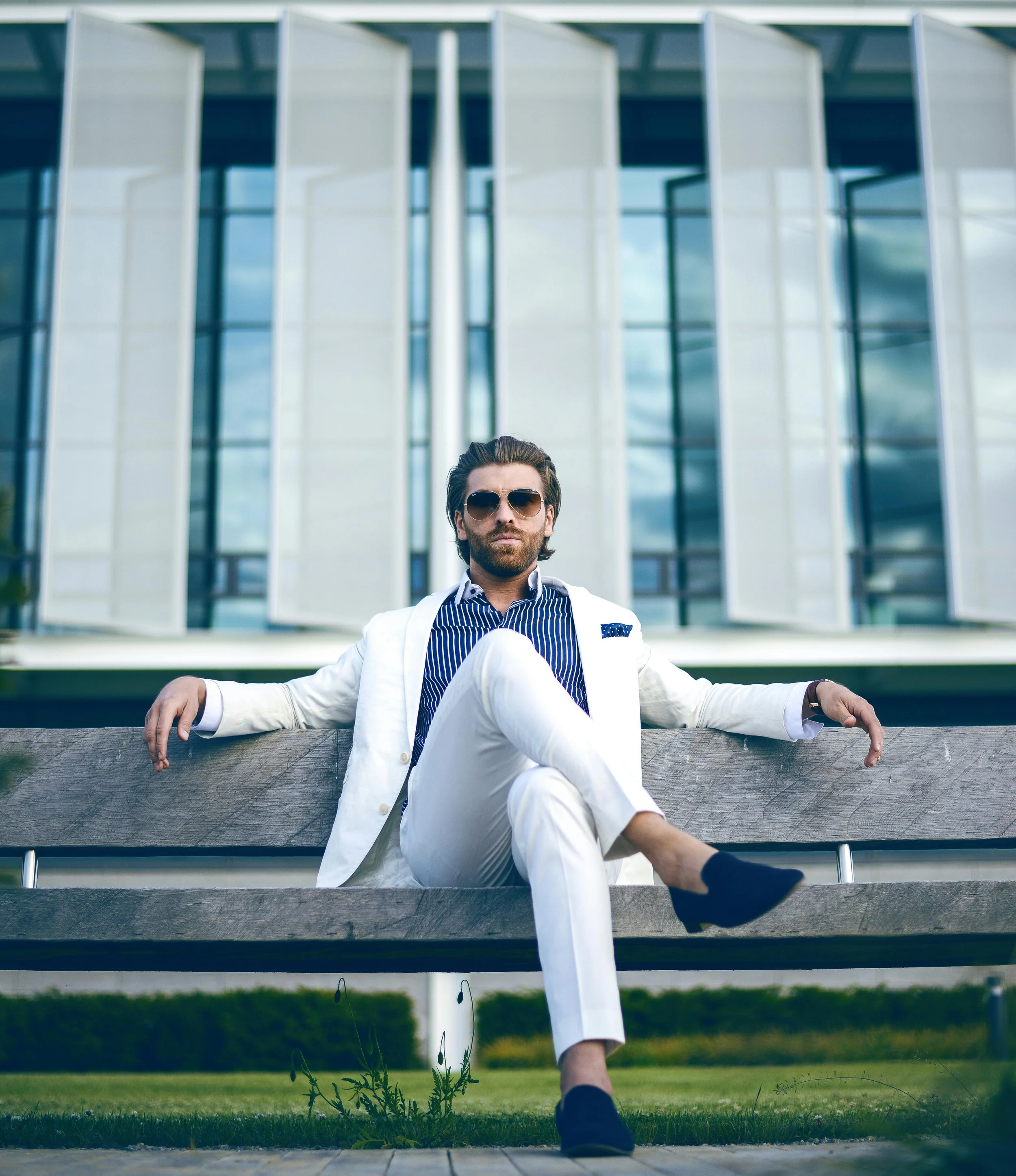 A man sitting on a bench in front of a modern glass building, wearing sunglasses and a white suit, with legs crossed and arms resting on the bench.