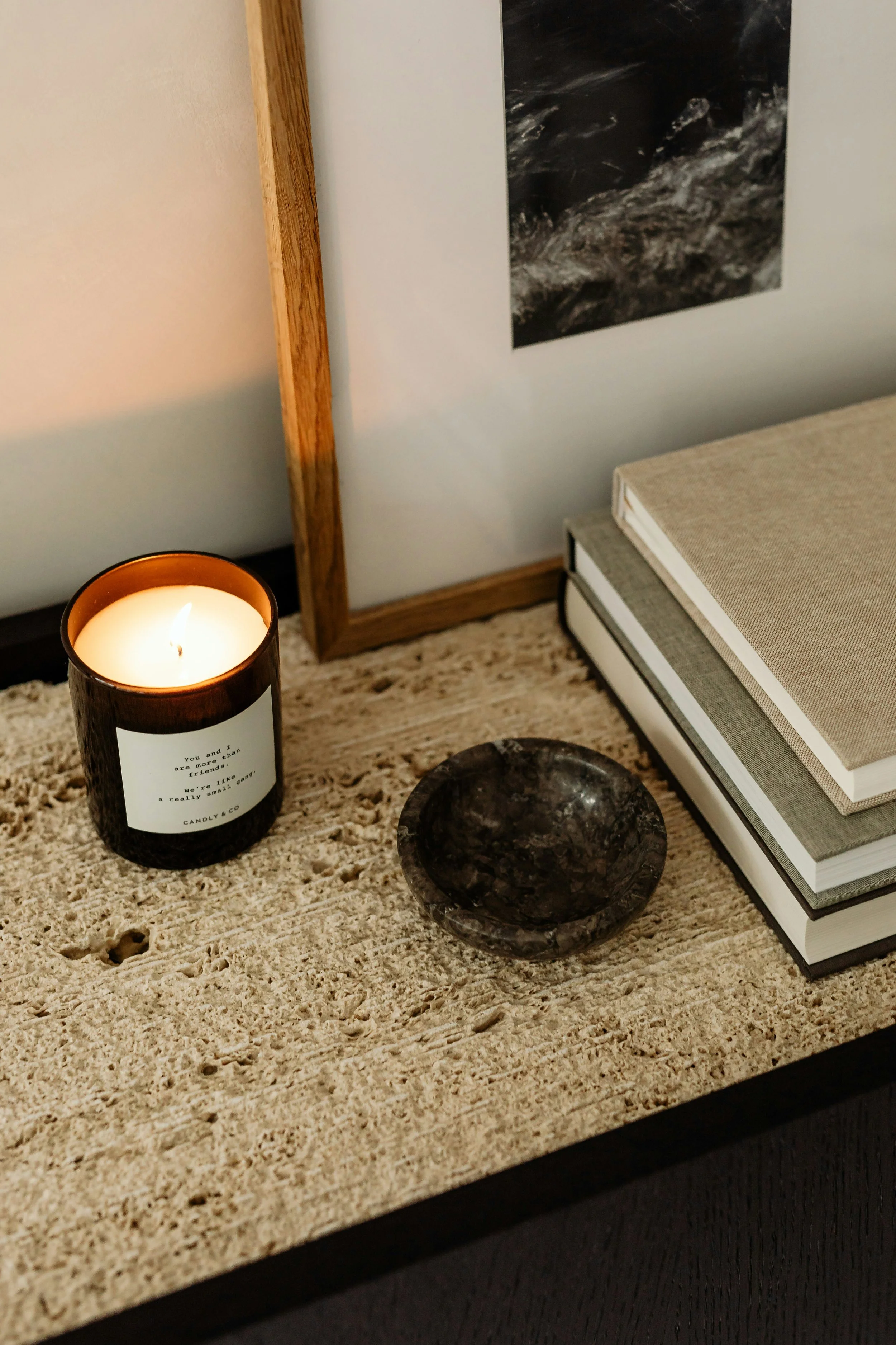 Close-up of a beige textured surface on a table with a lit candle, a dark marble sphere, and a stack of books. A part of a wooden frame and a black and white photo are visible in the background.
