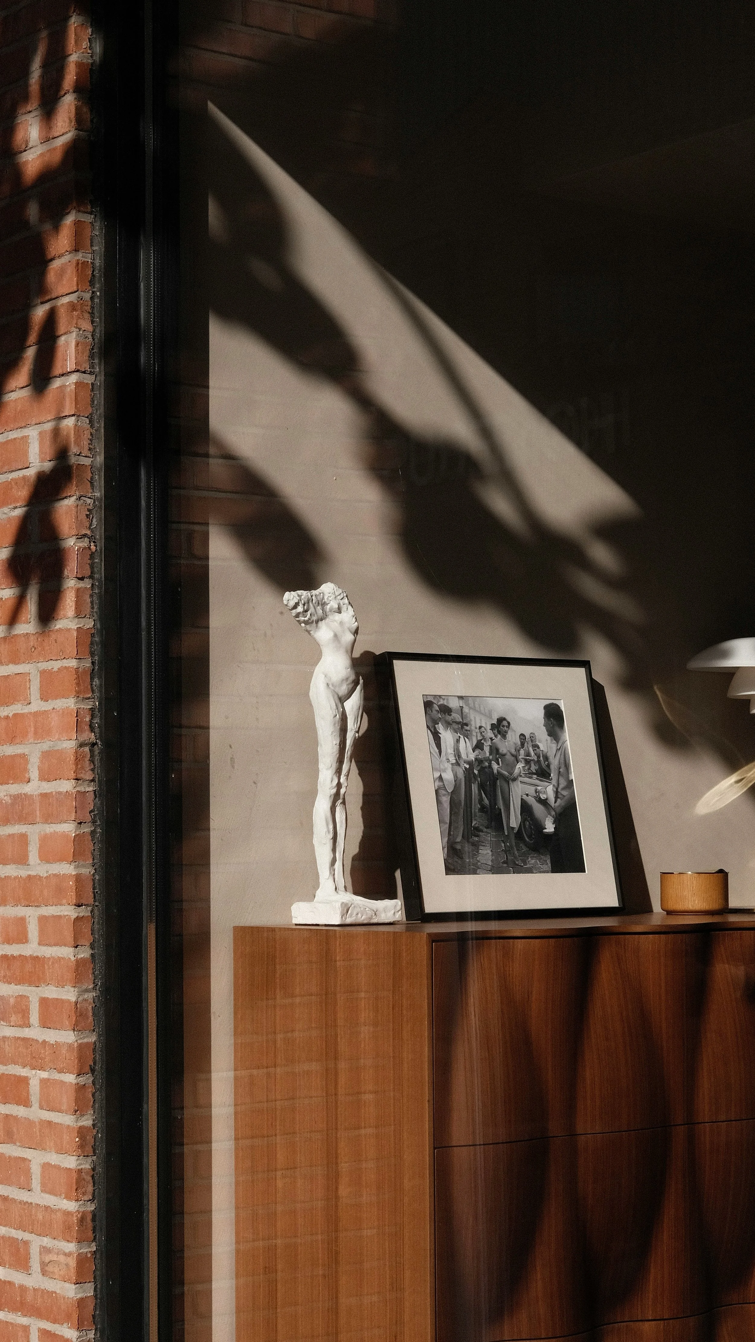 A decorative room corner with a white statue of a slender woman, a framed black-and-white photograph of people, and a small wooden bowl on a wooden cabinet against a wall with a brick and wood panel design. Shadows from nearby objects are cast on the wall.