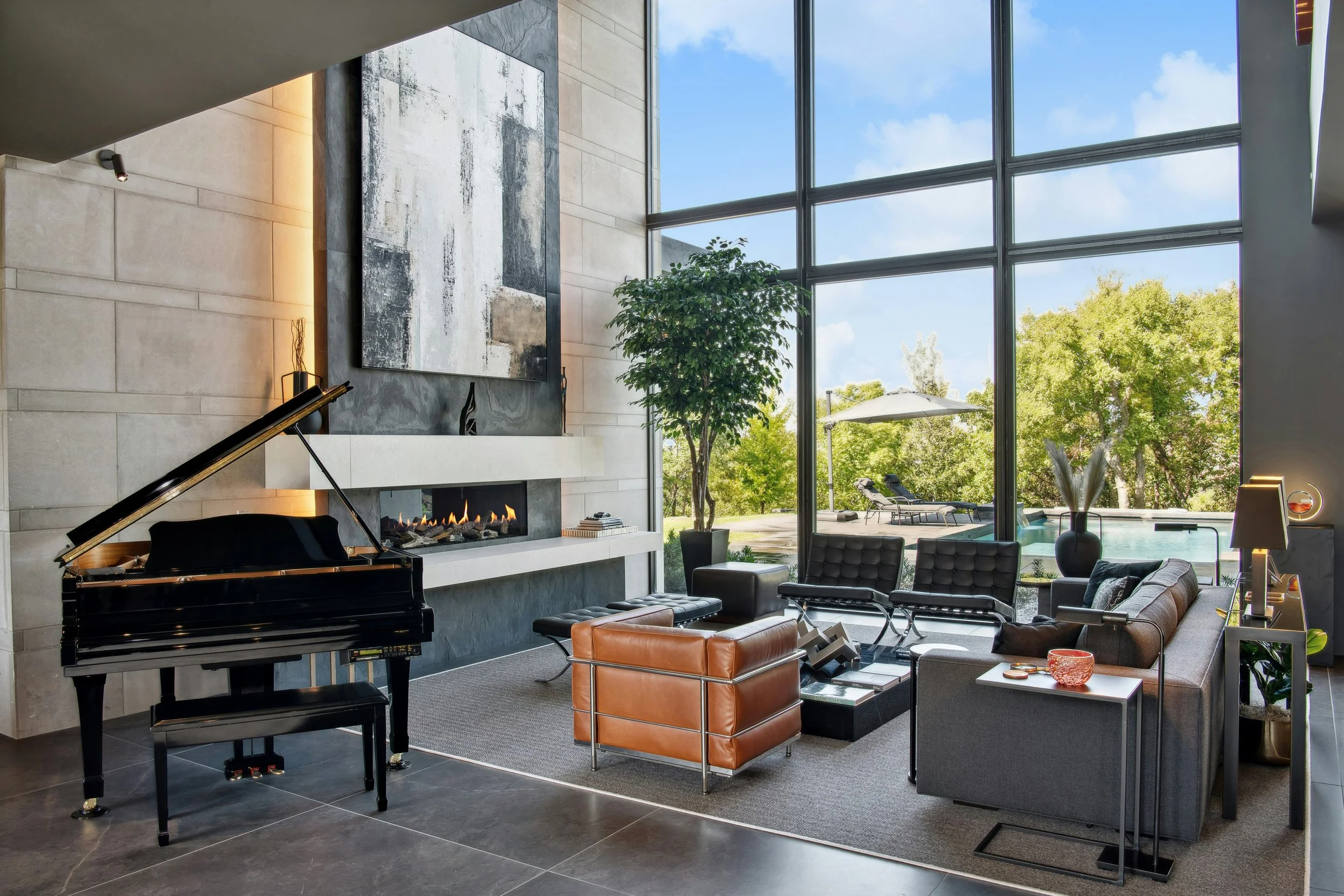 Modern living room with large floor-to-ceiling windows, a black grand piano, abstract artwork above a fireplace, and outdoor view of trees, pool, and lounge chairs.