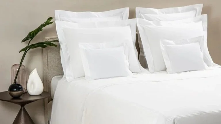 A neatly made bed with white sheets and pillows, a beige headboard, a bedside table with a glass vase holding a plant, and two decorative vases.