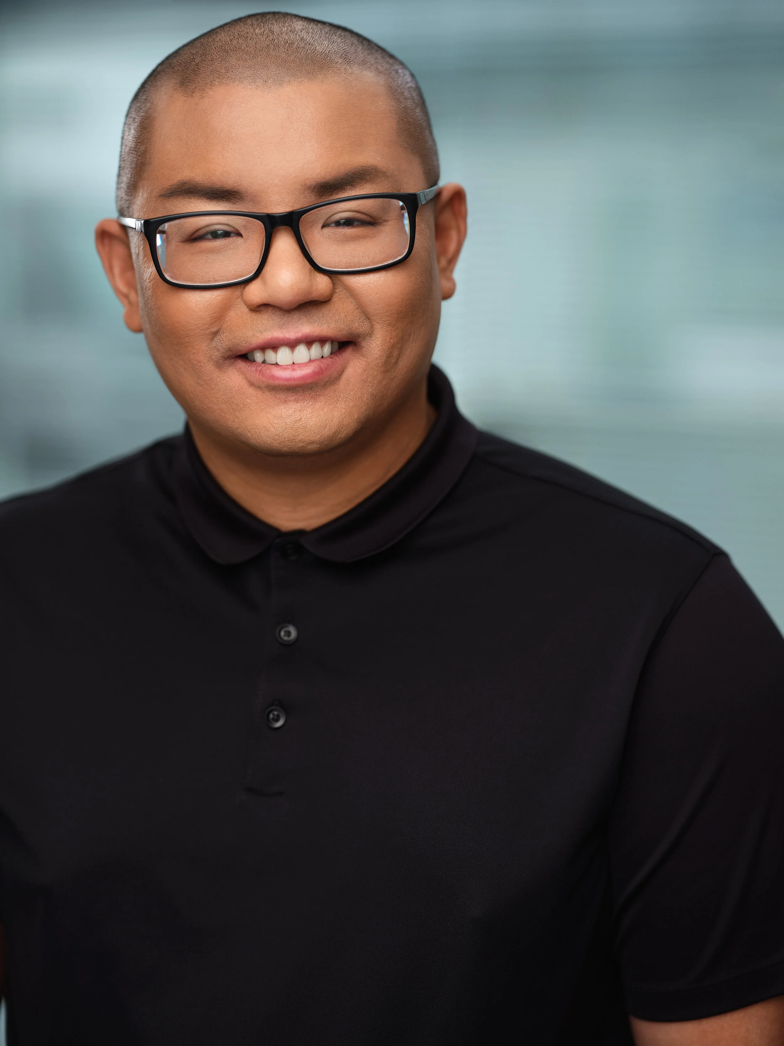 A smiling man with glasses, a shaved head, wearing a black polo shirt, against a blurred light blue background.