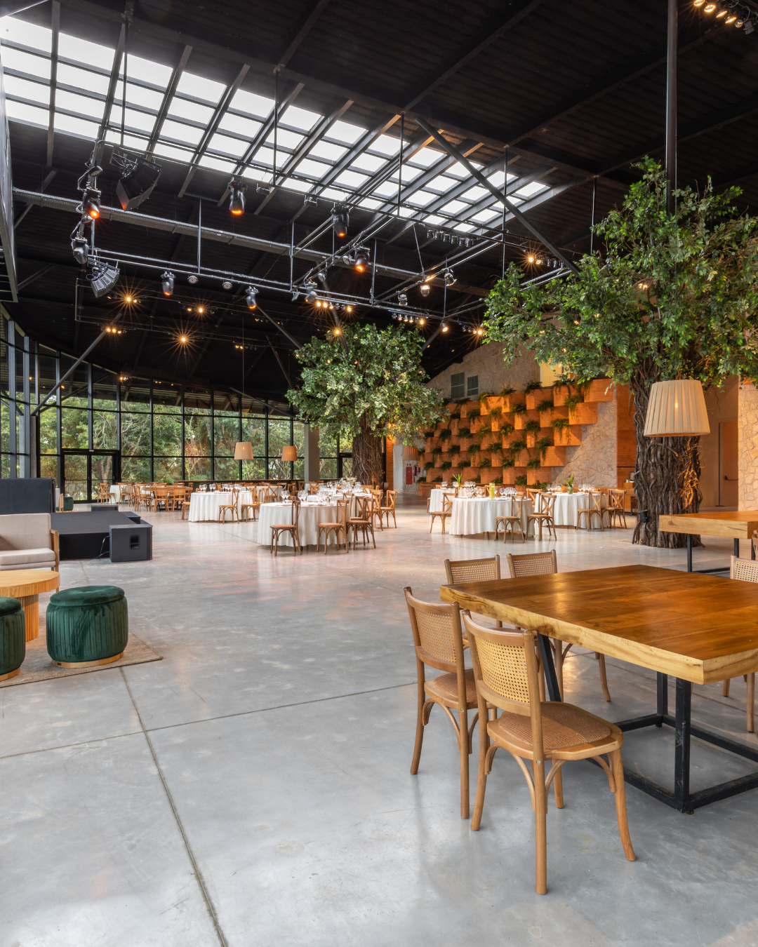 Elegant event space with round tables covered in white tablecloths, wooden chairs, large indoor trees, and a stage area in the background.