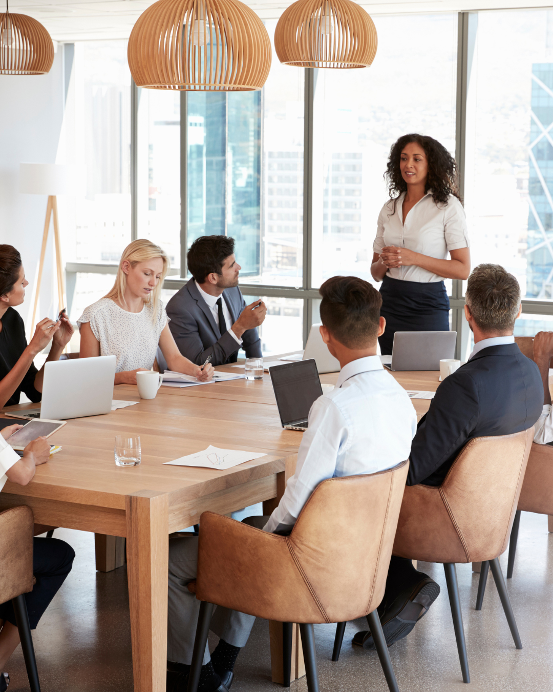 A woman in a white blouse and dark skirt speaks to a group in a modern office conference room with large windows, during a business meeting.