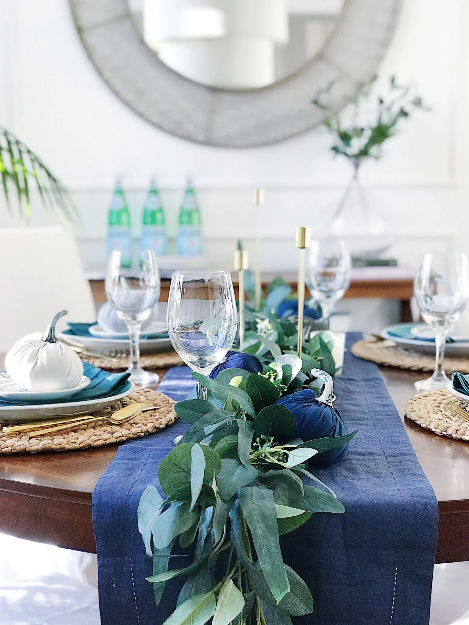 Dining table decorated for a meal with a blue table runner, woven placemats, wine glasses, plates with blue napkins, and a green garland centerpiece with small pumpkins and candles. In the background, there are bottles of sparkling water and a large mirror.