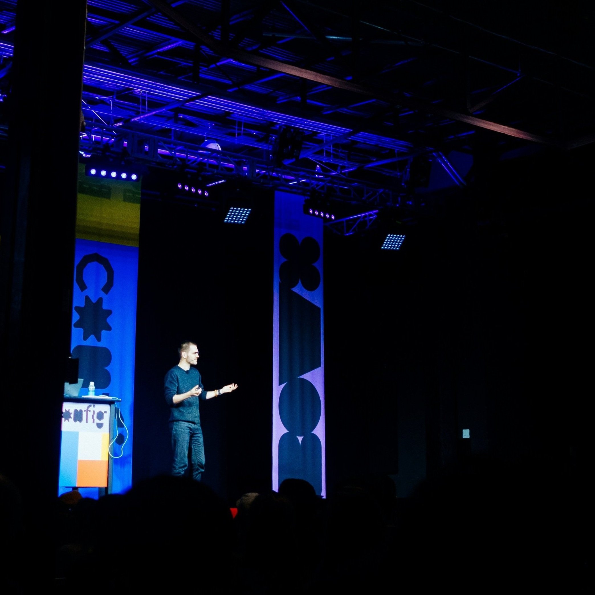 A man standing on a stage with a dark background, illuminated by blue stage lights, gesturing with his hands during a company presentation.