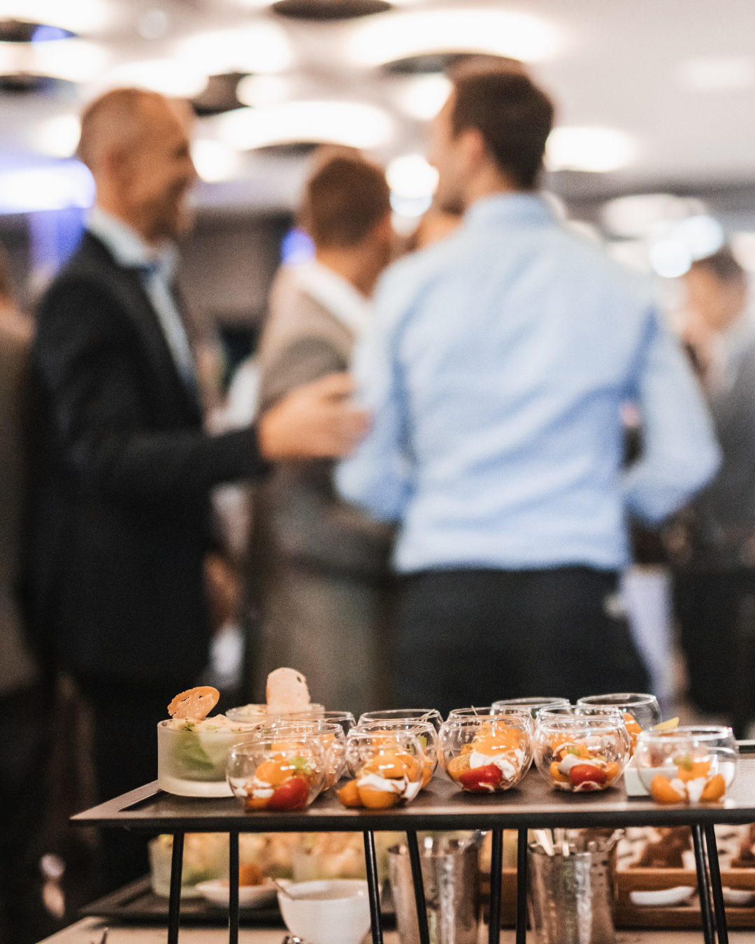 Appetizers and desserts on a tray at a social gathering with blurred people talking in the background.