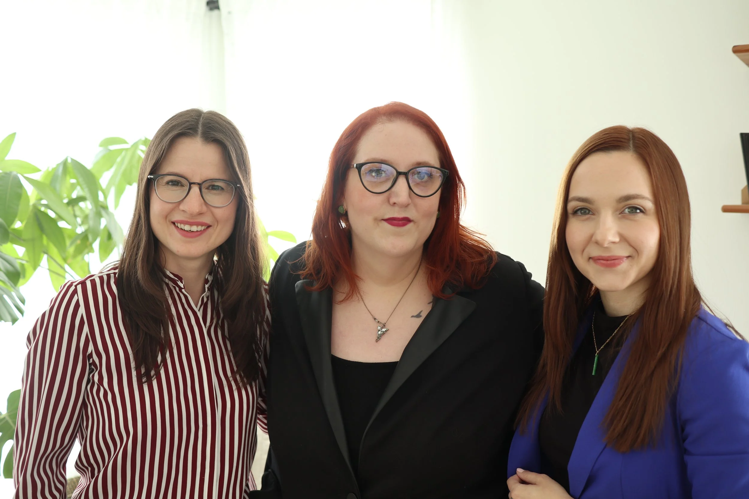 Three women with glasses standing close together indoors, smiling, with a leafy plant and window in the background.