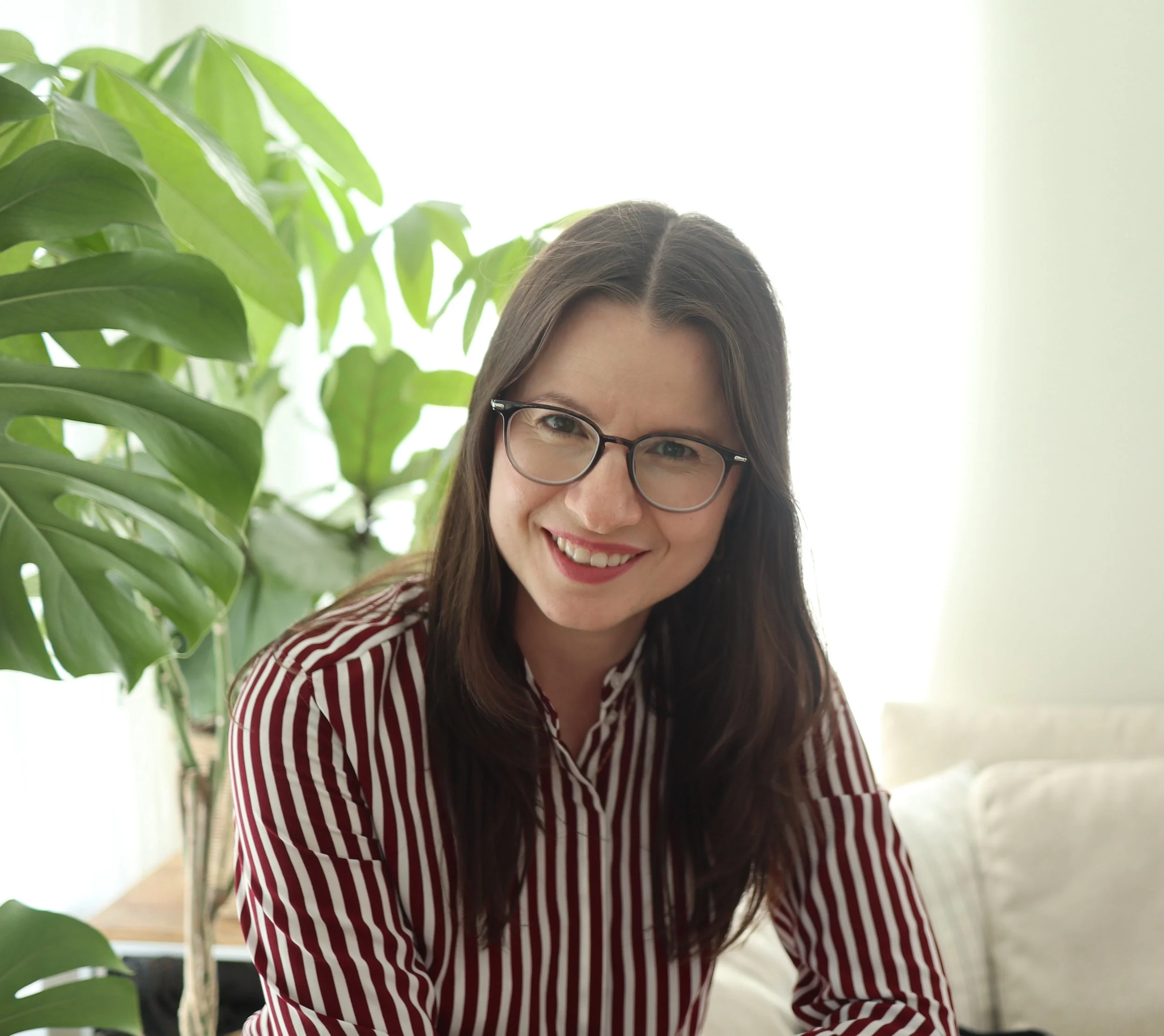 Smiling woman with glasses and long brown hair, wearing a red and white striped shirt, sitting near a large green plant in a bright room.