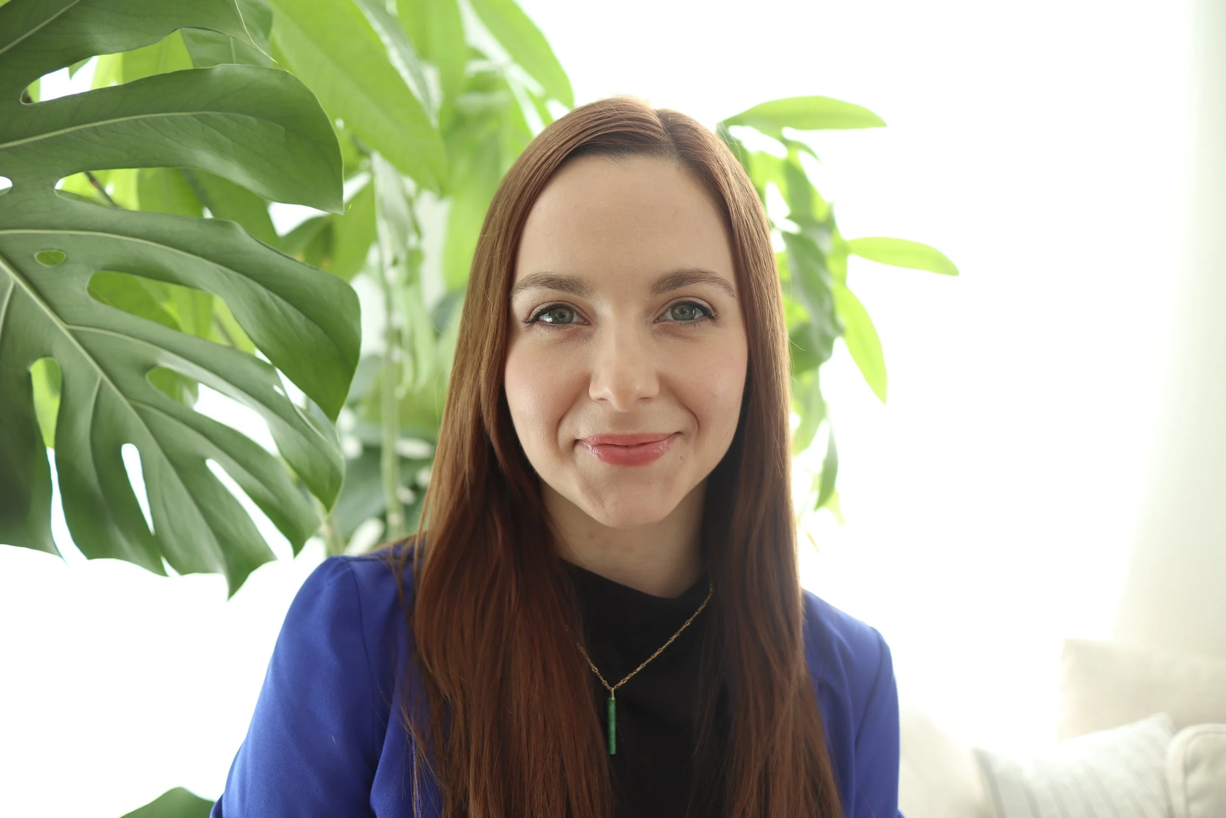 A woman with long, straight, reddish-brown hair smiling at the camera, wearing a blue blazer and a gold necklace with a green pendant, in front of green houseplants.