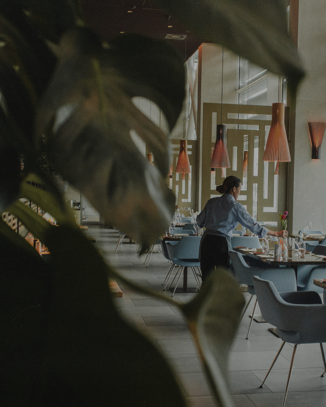 A waitress preparing a table in a modern restaurant with blue chairs, wooden dividers, pink pendant lights, and large windows.
